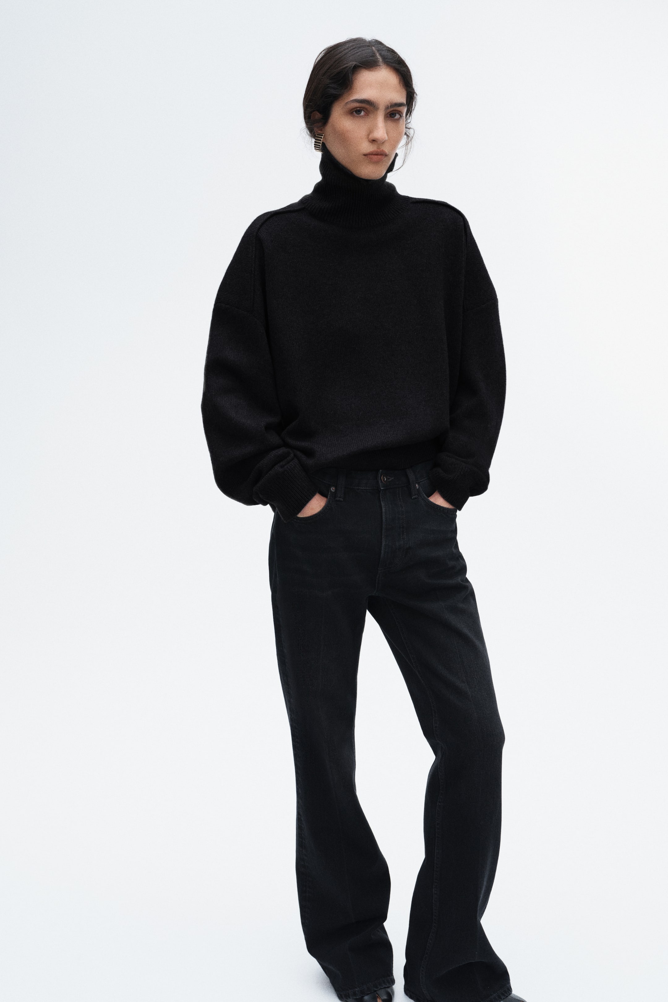 Grösseres Bild ansehen: Ein schwarzer Oversize-Rollkragenpullover mit überschnittenen Schultern und Rippbündchen wird vom Model getragen. Das Model trägt ausserdem schwarze High-Waist-Jeans mit geradem Bein und leichtem Schlag und hat die Hände in den vorderen Taschen.