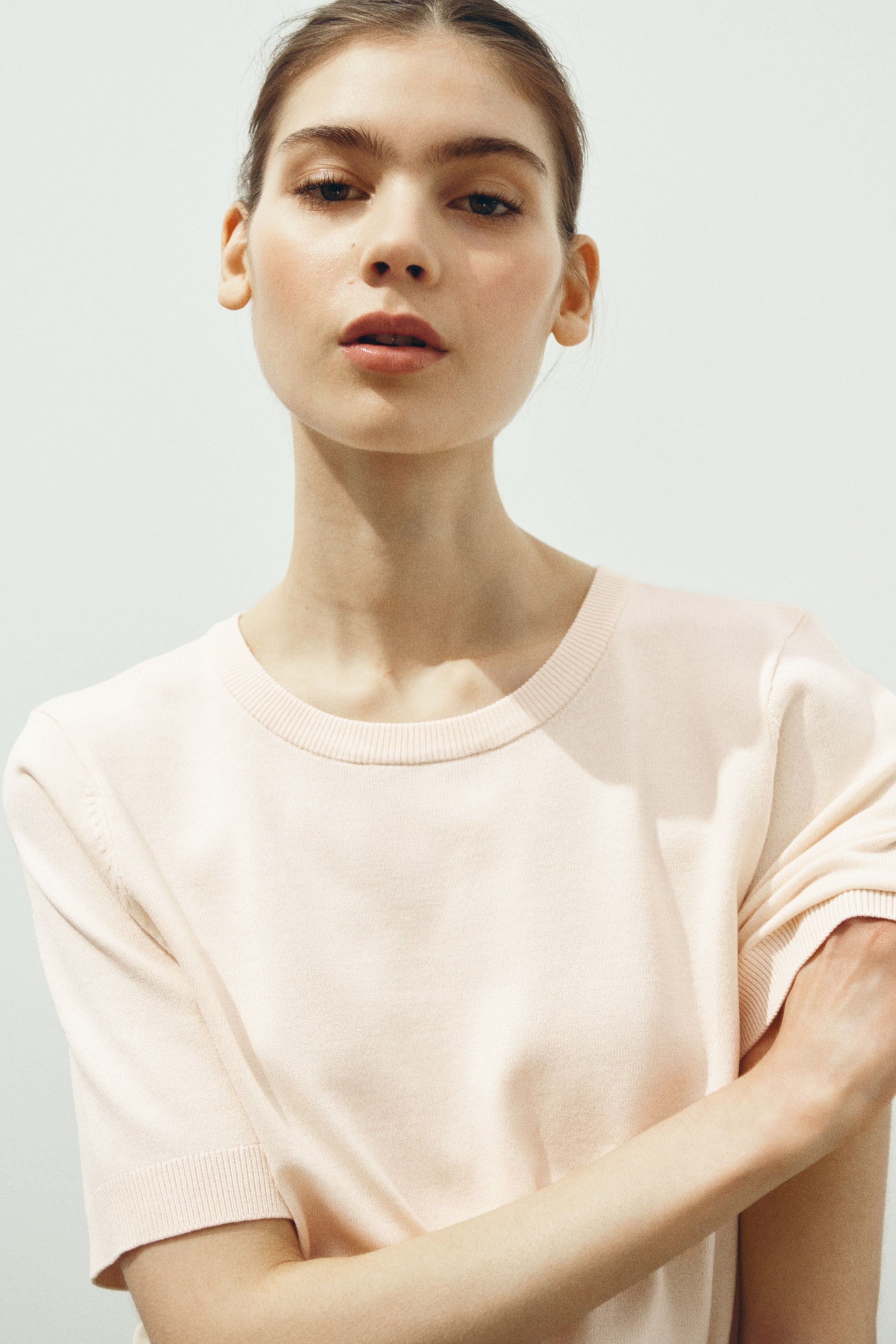 查看大圖: 模特穿著一件淺粉色細針織短袖上衣，領口和袖口具有羅紋設計。模特雙臂交叉。