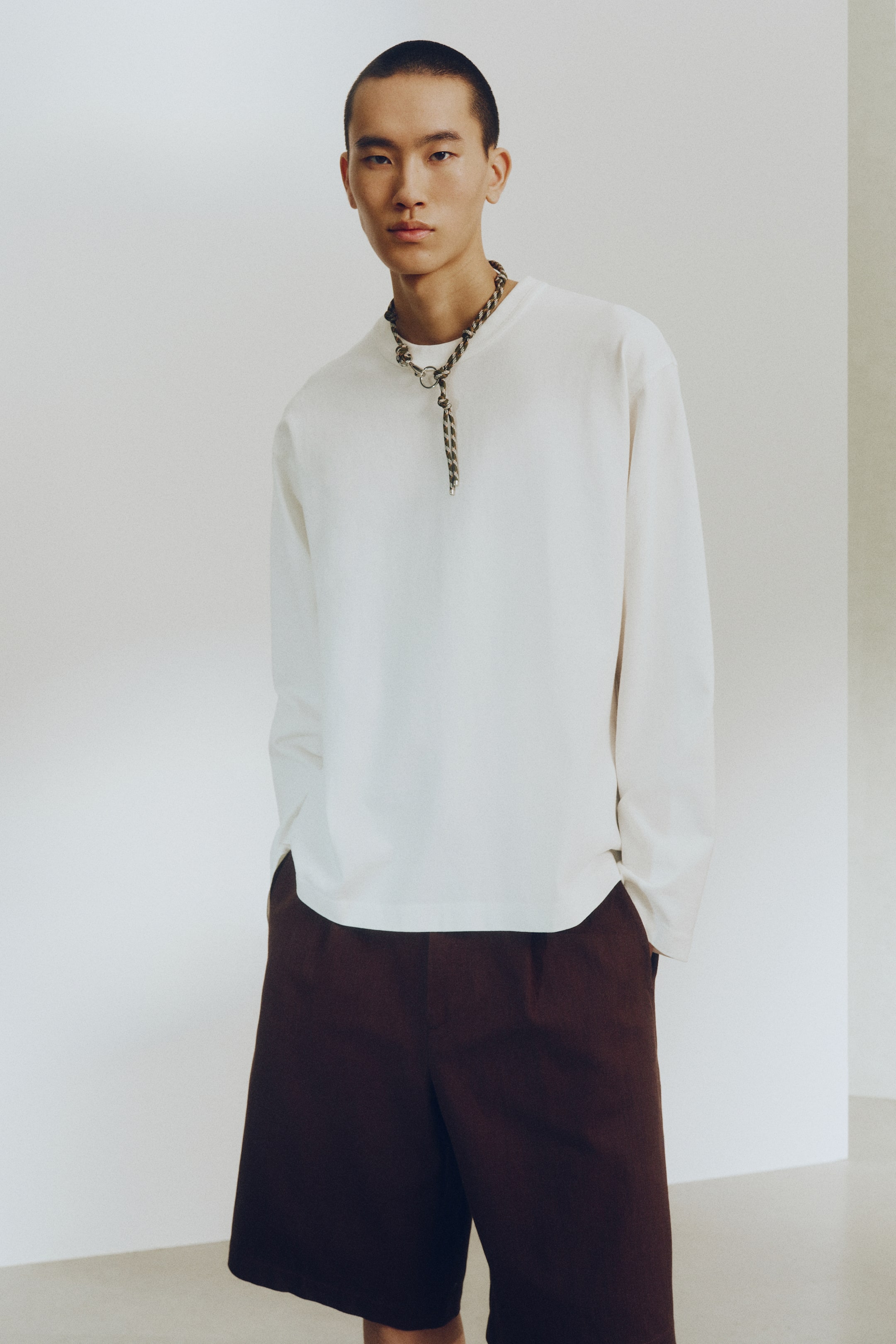 View larger image: A white long-sleeve crew neck t-shirt with a relaxed fit is paired with dark brown relaxed-fit shorts. The look is completed with a braided necklace featuring a metal ring and toggle closure, elegantly resting on the t-shirt.