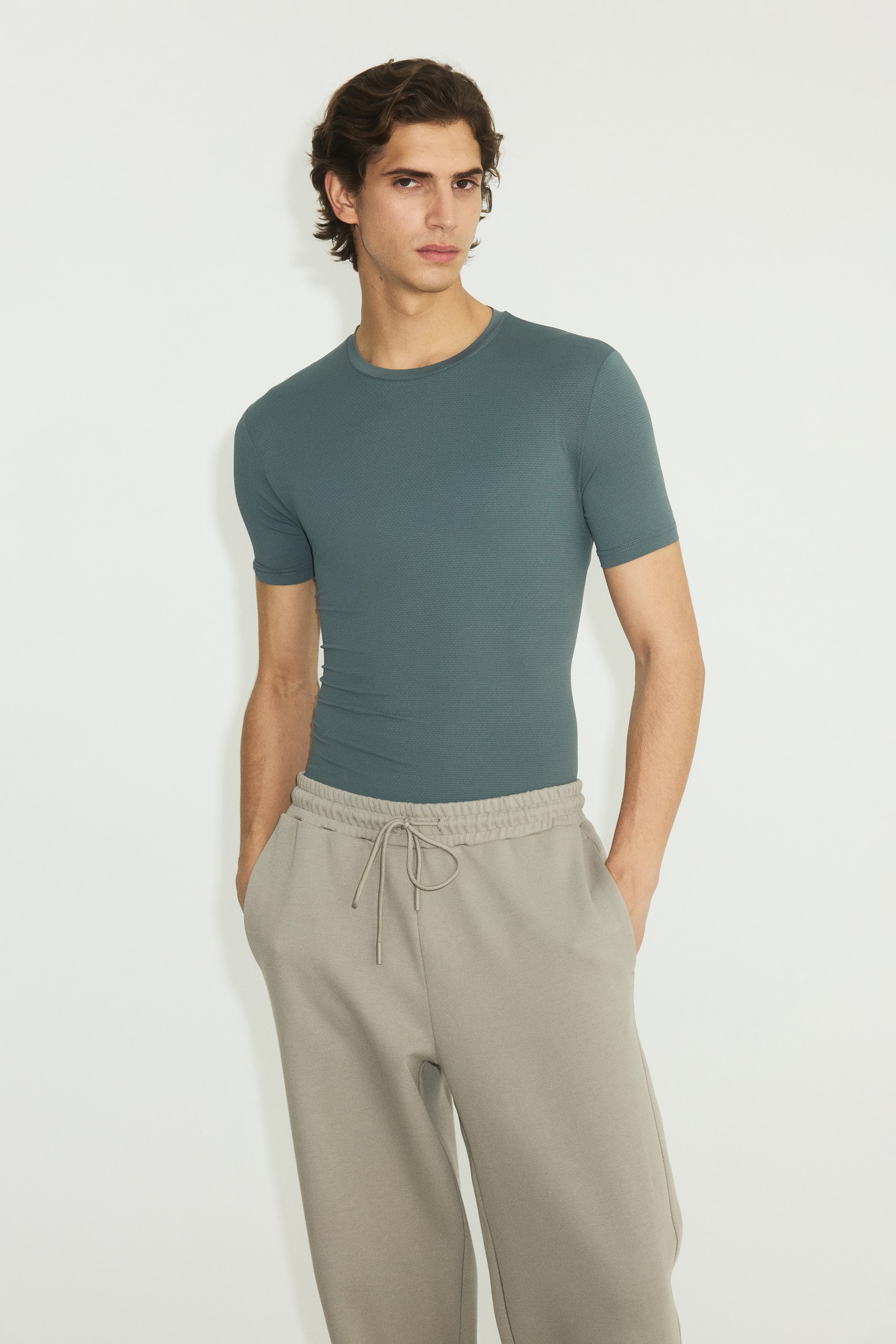Büyük resmi görüntüle: Koyu teal, kısa kollu, slim-fit tişört, ince dokulu bir desen ve bisiklet yaka ile tasarlanmış olup, elastik bağcıklı bel bandı ve yan cepleri bulunan rahat kesim açık gri eşofman altına sokulmuş şekilde giyilmektedir.