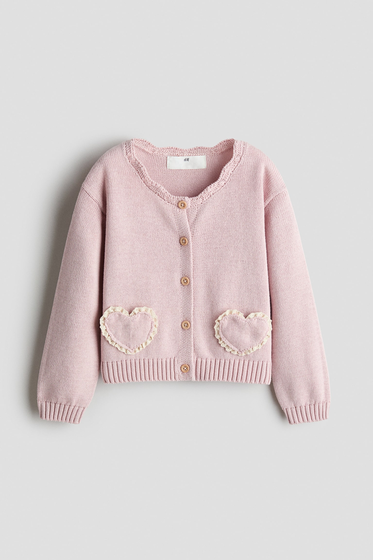 Eine hellrosa Strickjacke ist von vorne zu sehen, mit rundem Ausschnitt und gewellter Borte sowie einer FÃ¼nf-Knopf-Schliessung in der Mitte. Sie hat lange Ãrmel mit gerippten BÃ¼ndchen, einen gerippten Saum und zwei aufgesetzte Taschen vorne in Herzform mit cremefarbenen, gerÃ¼schten SpitzenrÃ¤ndern.