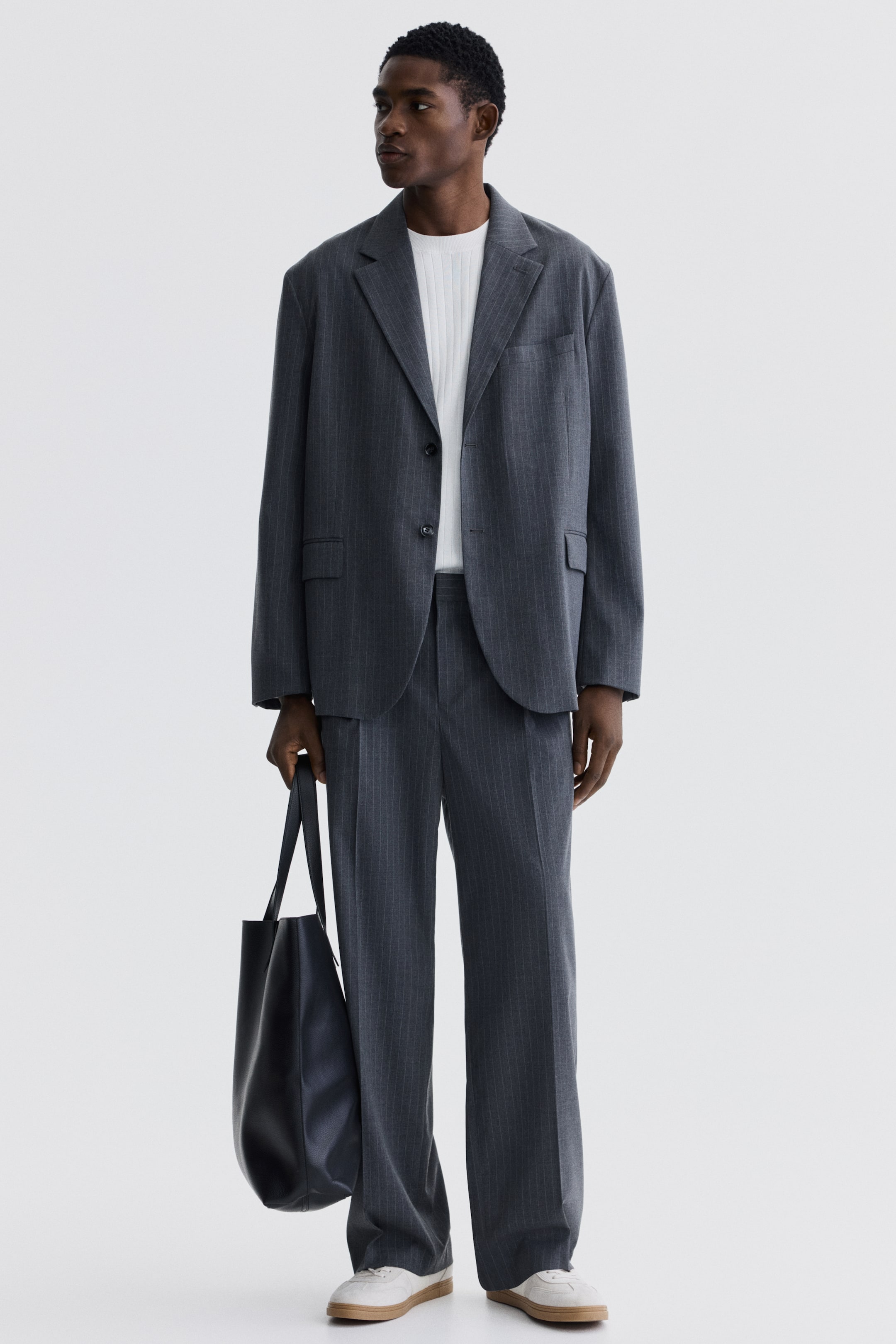 拡大画像を表示: モデルが着用しているのは、ダークグレーのストライプ入りテーラードパンツです。ゆったりとしたストレートレッグフィットで、目立つ折り目が特徴です。さらに、モデルはそれに合わせたジャケットも着用しています。