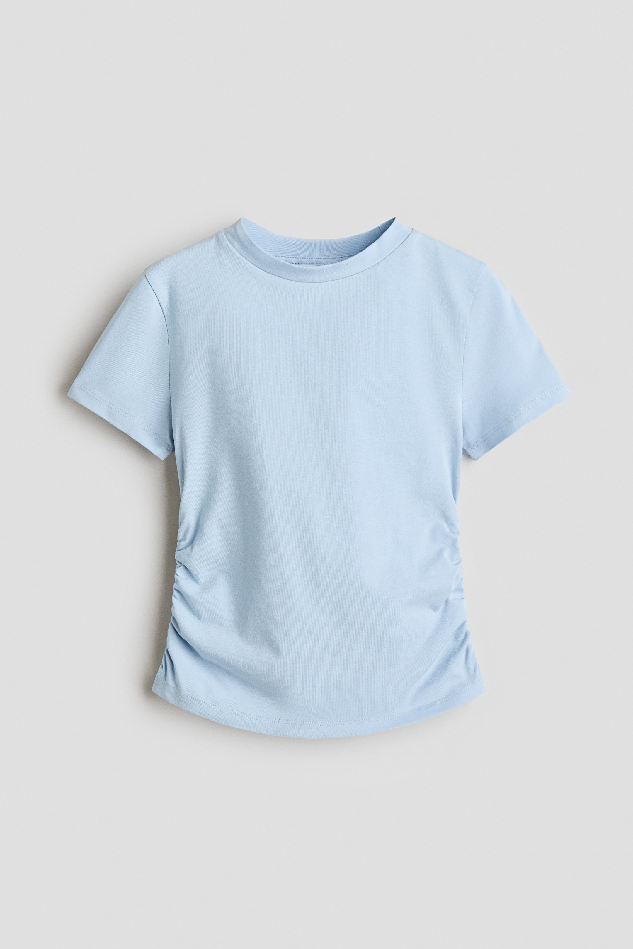 Grotere afbeelding bekijken: Een lichtblauw T-shirt, van voren afgebeeld, met een ronde hals, korte mouwen en gerimpelde details langs de onderste zijnaden die een geplooid effect creëren.