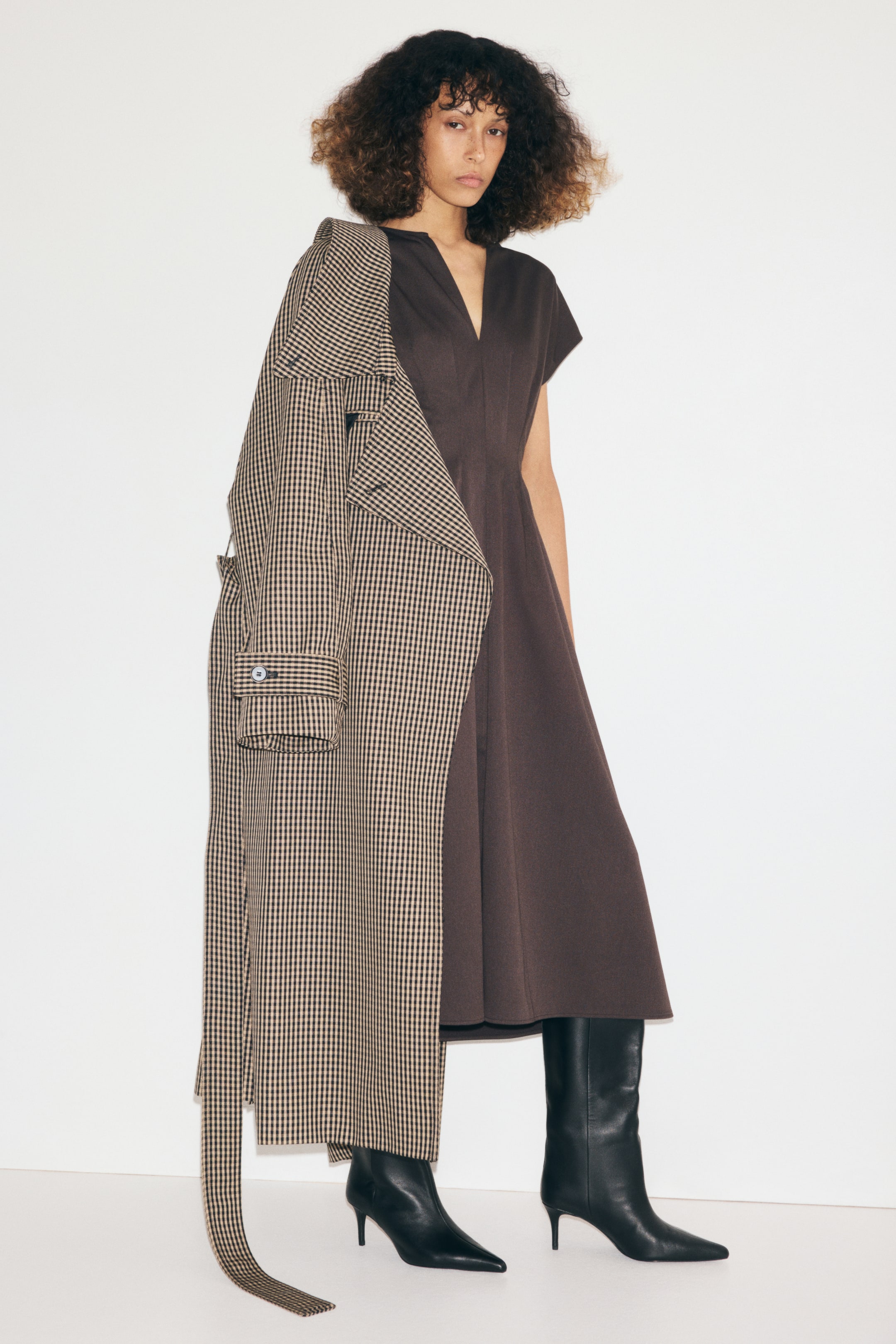 Ampliar la imagen: La modelo lleva un vestido color café oscuro, a la pantorrilla, con escote en V, mangas cortas y falda evasé. Sobre un hombro del vestido lleva un trench coat a cuadros en tonos café y beige, combinado con botas negras de cuero con tacón alto.