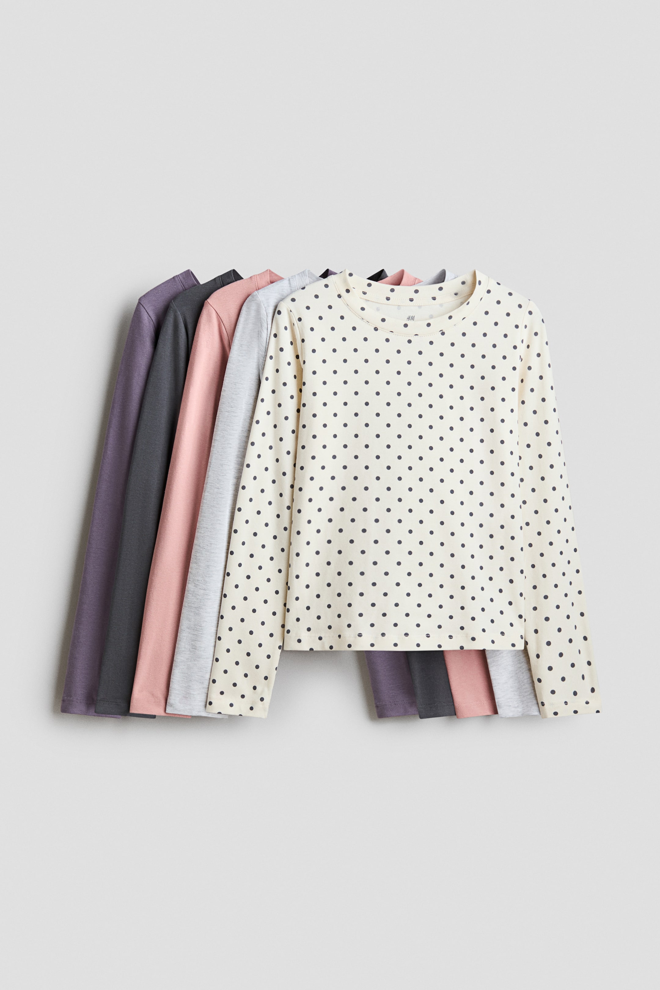 Visa större bild: Långärmade T-shirts i mjuk bomullsjersey visas upp, där den främsta tröjan har en krämfärgad bakgrund med mörkgrå prickar och en rund halsringning med smal kant. Flera enfärgade långärmade T-shirts ligger lager på lager bakom den, inklusive ljusgrå, rosa, mörkgrå och lila.