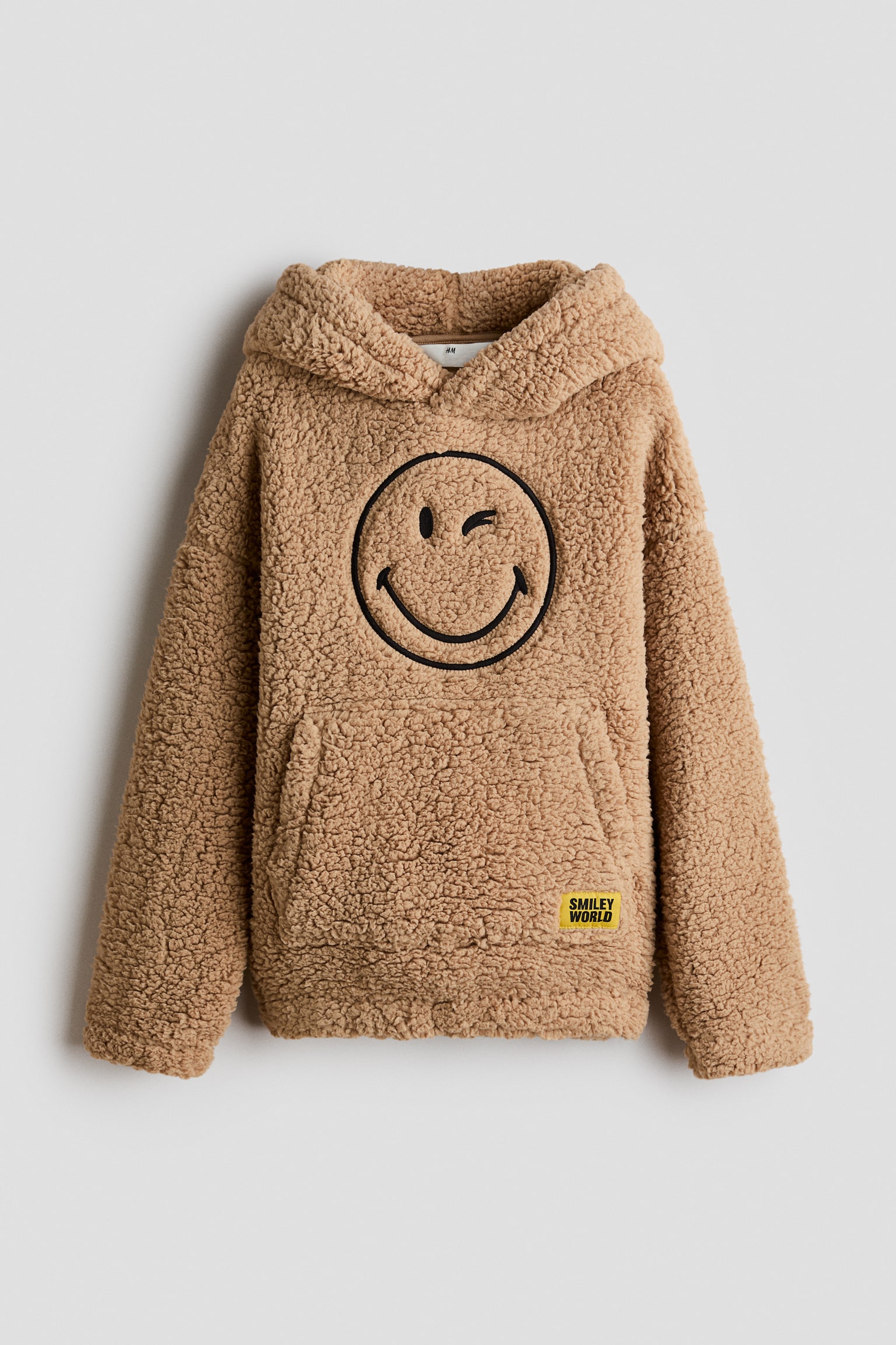 Sveta braon teddy fleece duks sa kapuljaÄom prikazan je spreda, sa velikim crnim izvezenim namigujucÌim smajlijem na grudima. Dizajn prikazuje kapuljaÄu sa preklopom napred, spuÅ¡tena ramena, elastiÄne manÅ¾etne i kengur dÅ¾ep sa malom Å¾utom logo zakrpom.
