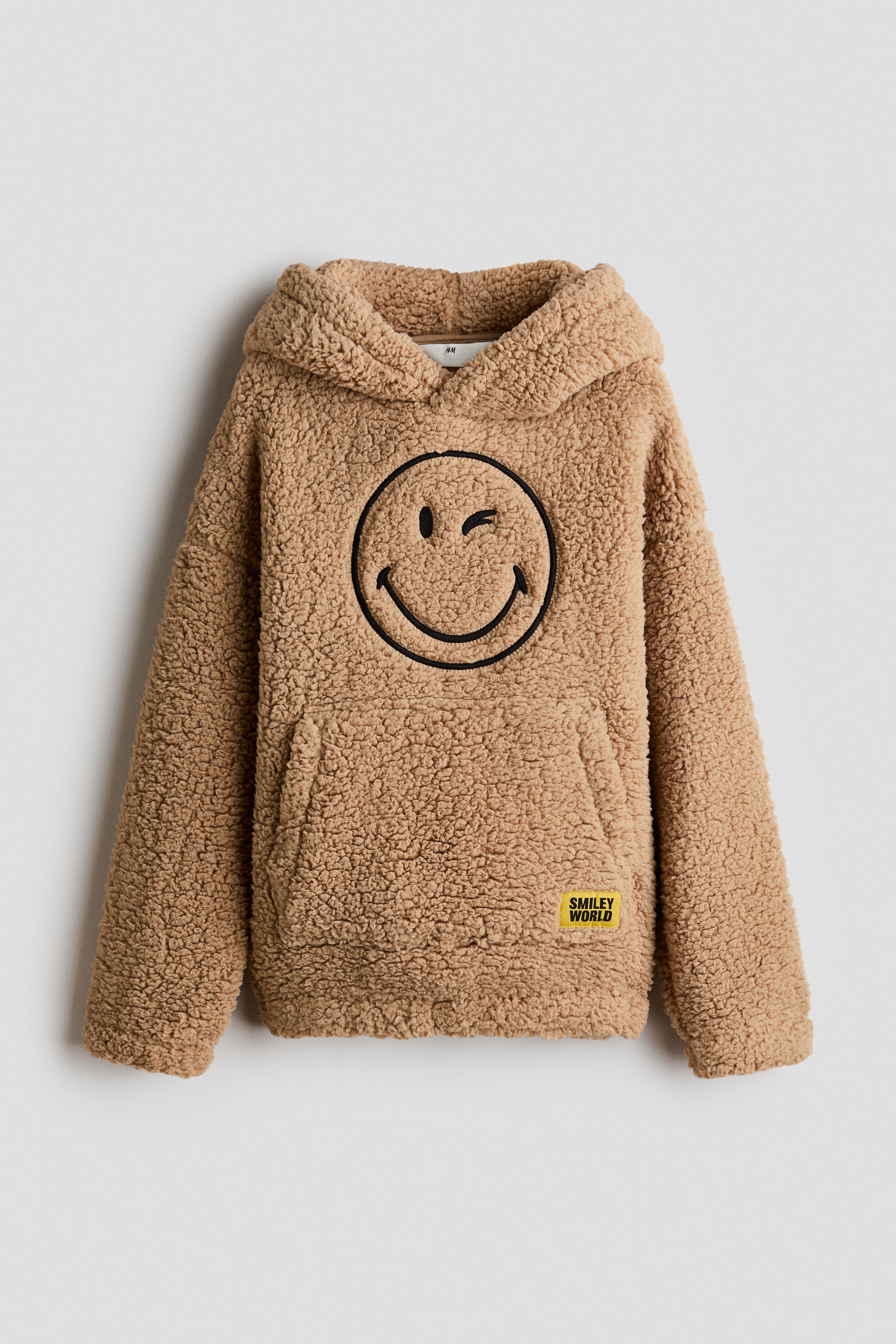 Prikaži veću sliku: Sveta braon teddy fleece duks sa kapuljačom prikazan je spreda, sa velikim crnim izvezenim namigujućim smajlijem na grudima. Dizajn prikazuje kapuljaču sa preklopom napred, spuštena ramena, elastične manžetne i kengur džep sa malom žutom logo zakrpom.