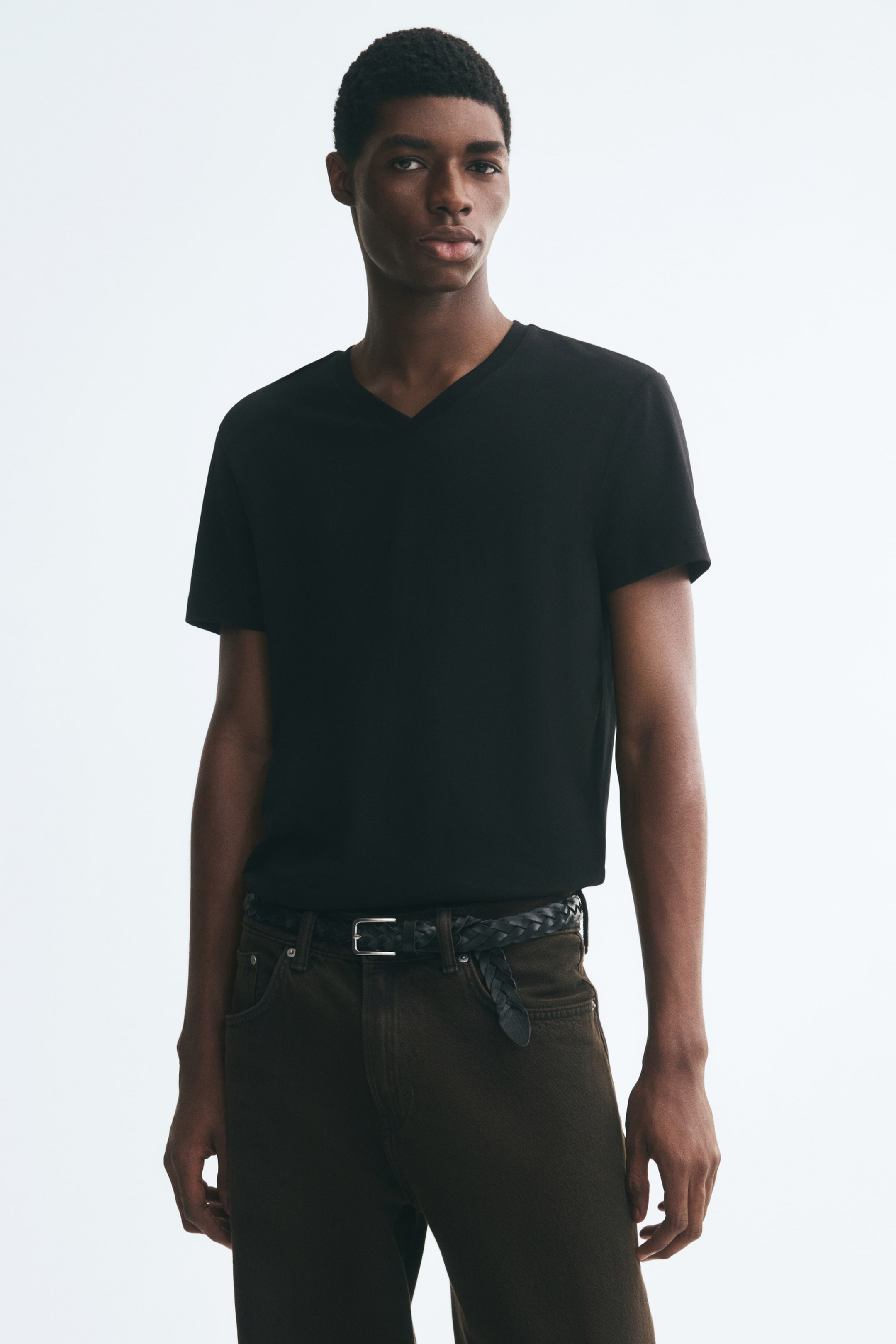 Größeres Bild anzeigen: Ein schwarzes, schmal geschnittenes T-Shirt mit V-Ausschnitt wird vom Model getragen und in dunkelbraune Hose gesteckt, die mit einem schwarzen geflochtenen Gürtel fixiert ist. Das leichte Baumwoll-Jersey-Shirt verfügt über kurze Ärmel und eine figurbetonte Silhouette.