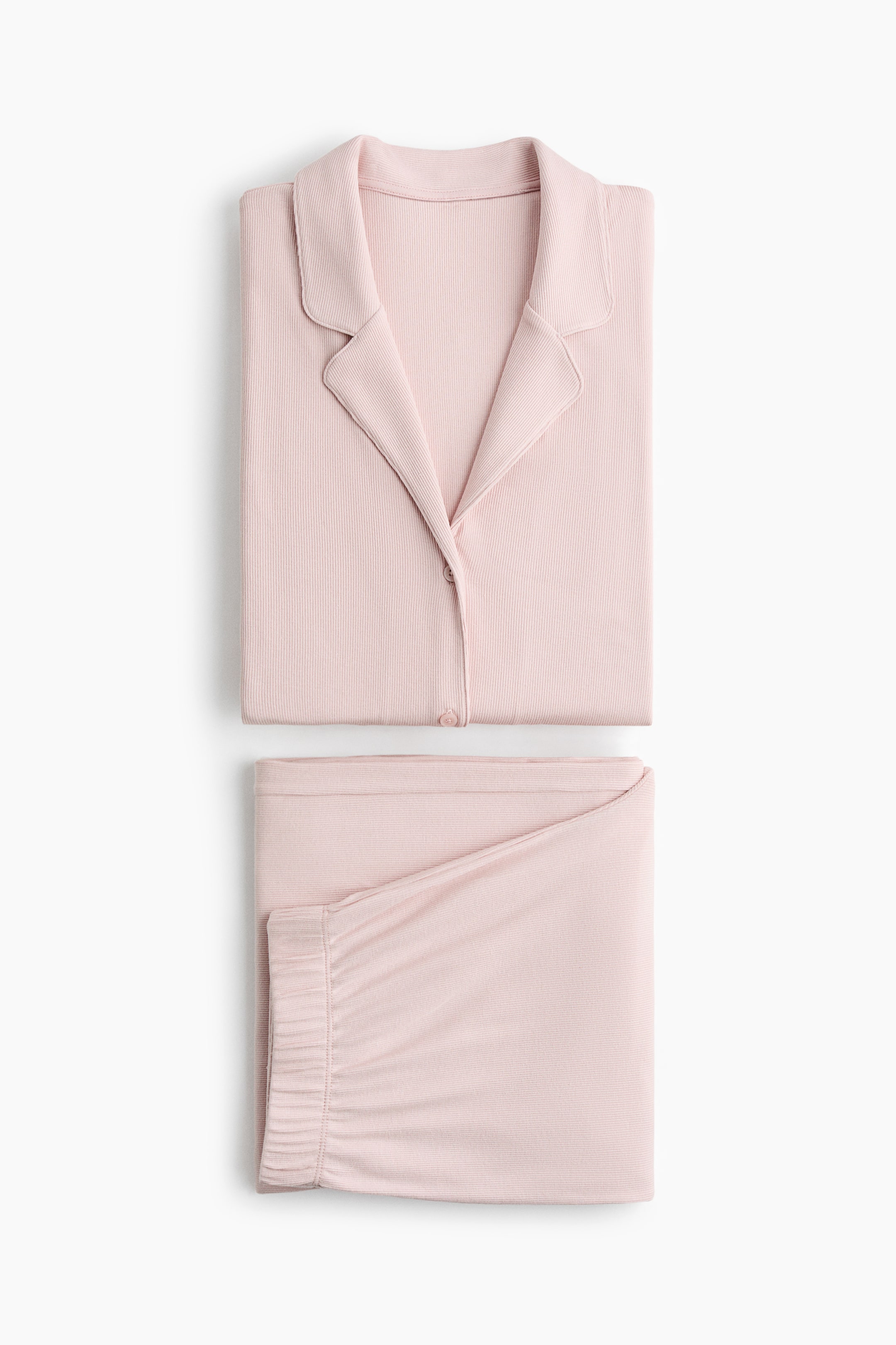 Nagyobb kép megtekintése: Egy rózsaszín bordázott pizsama szett látható, amely egy hajtogatott felsőt és a hozzá illő hajtogatott nadrágot tartalmaz. A felső, amelyet elölről mutatnak be, gallérral és látható gombos előlappal rendelkezik, míg a nadrág összehúzott, rugalmas derékpánttal rendelkezik.