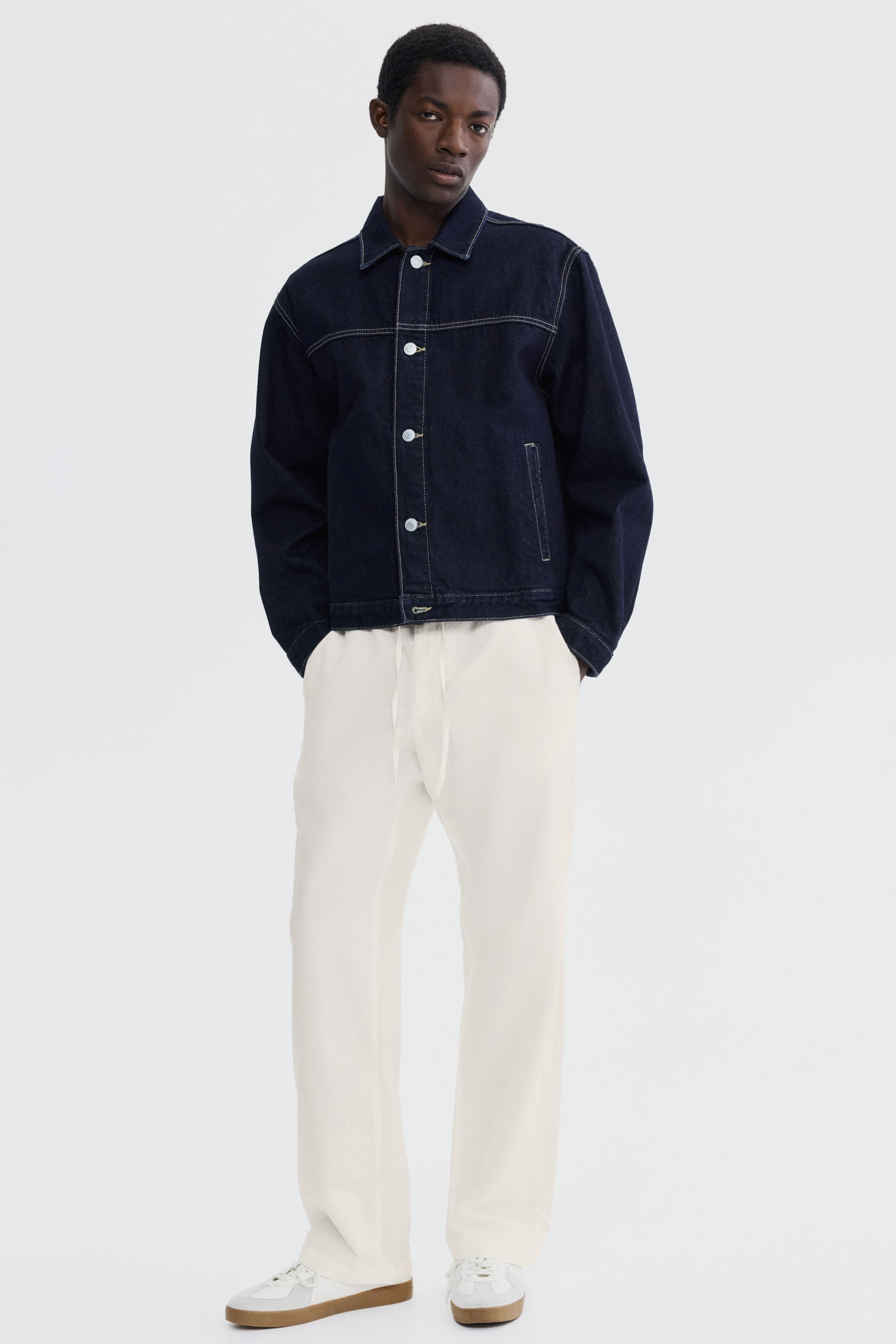 View larger image: A dark wash denim jacket features white contrast stitching, a pointed collar, and white buttons down the front placket, with a patch pocket on the left side, presenting a relaxed fit. It's styled with cream-colored, wide-leg drawstring pants and white low-top sneakers featuring light gray accents and gum soles, all facing forward.