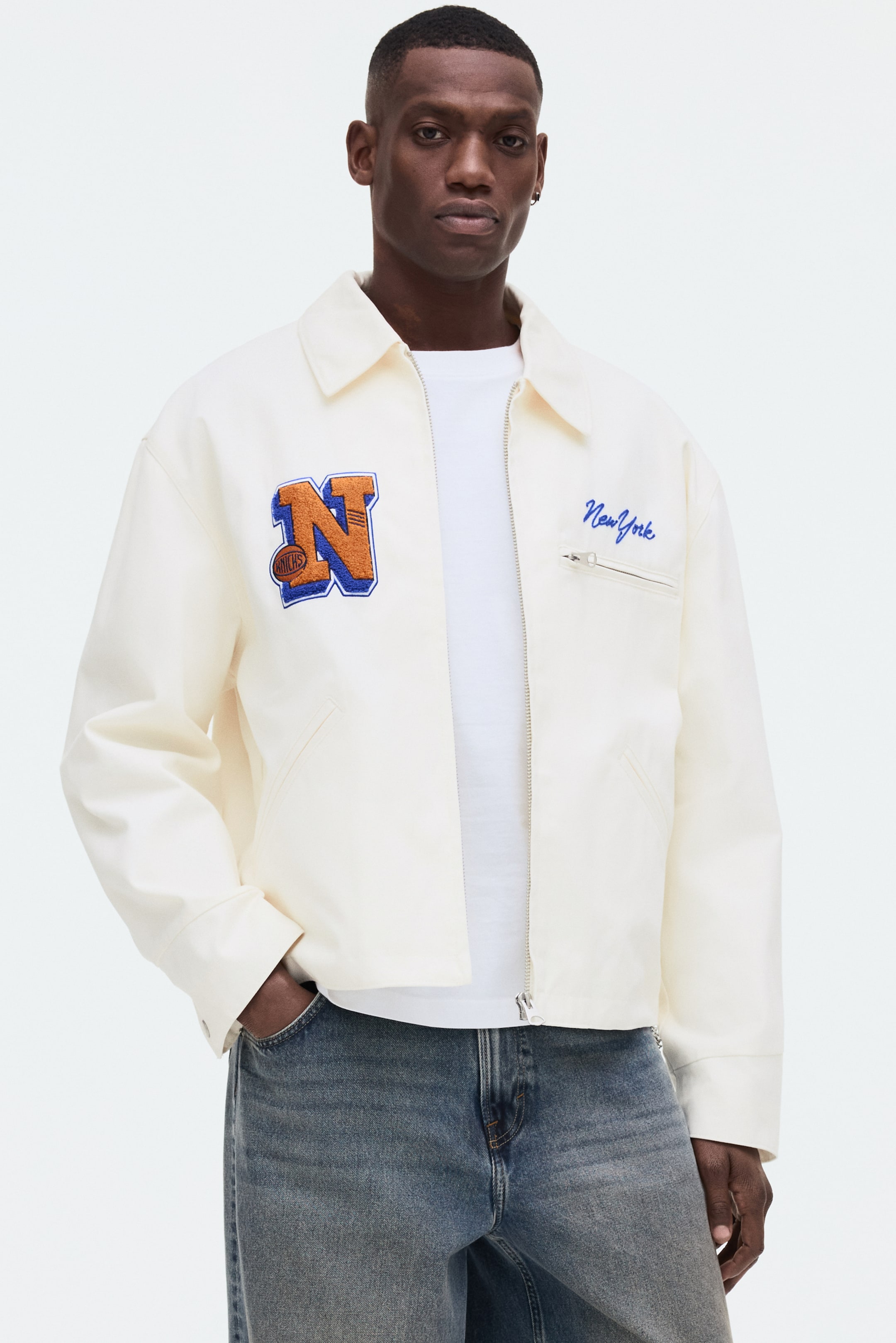 View larger image: The cream-colored jacket showcases a full-length silver zipper and a pointed collar, styled open over a white t-shirt. It features an orange and blue 'N' patch with a basketball graphic on the left chest, blue 'New York' script embroidery on the right chest, two angled welt pockets, and a zippered chest pocket.