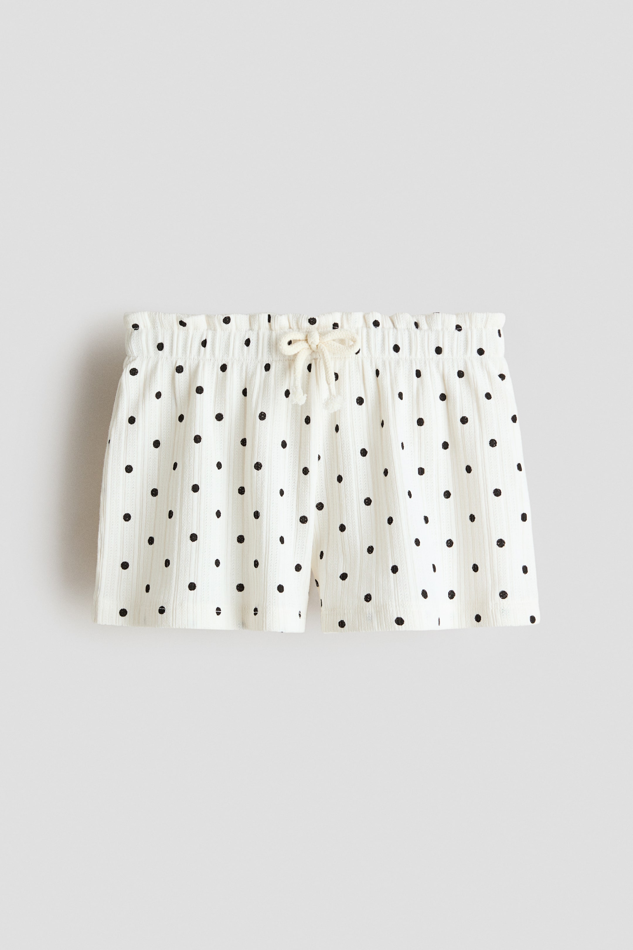 View larger image: The shorts are showcased from the front, crafted from an off-white pointelle jersey fabric adorned with black polka dots. They come with an elasticated, gathered waistband, complemented by a light-coloured drawstring tied in a bow.
