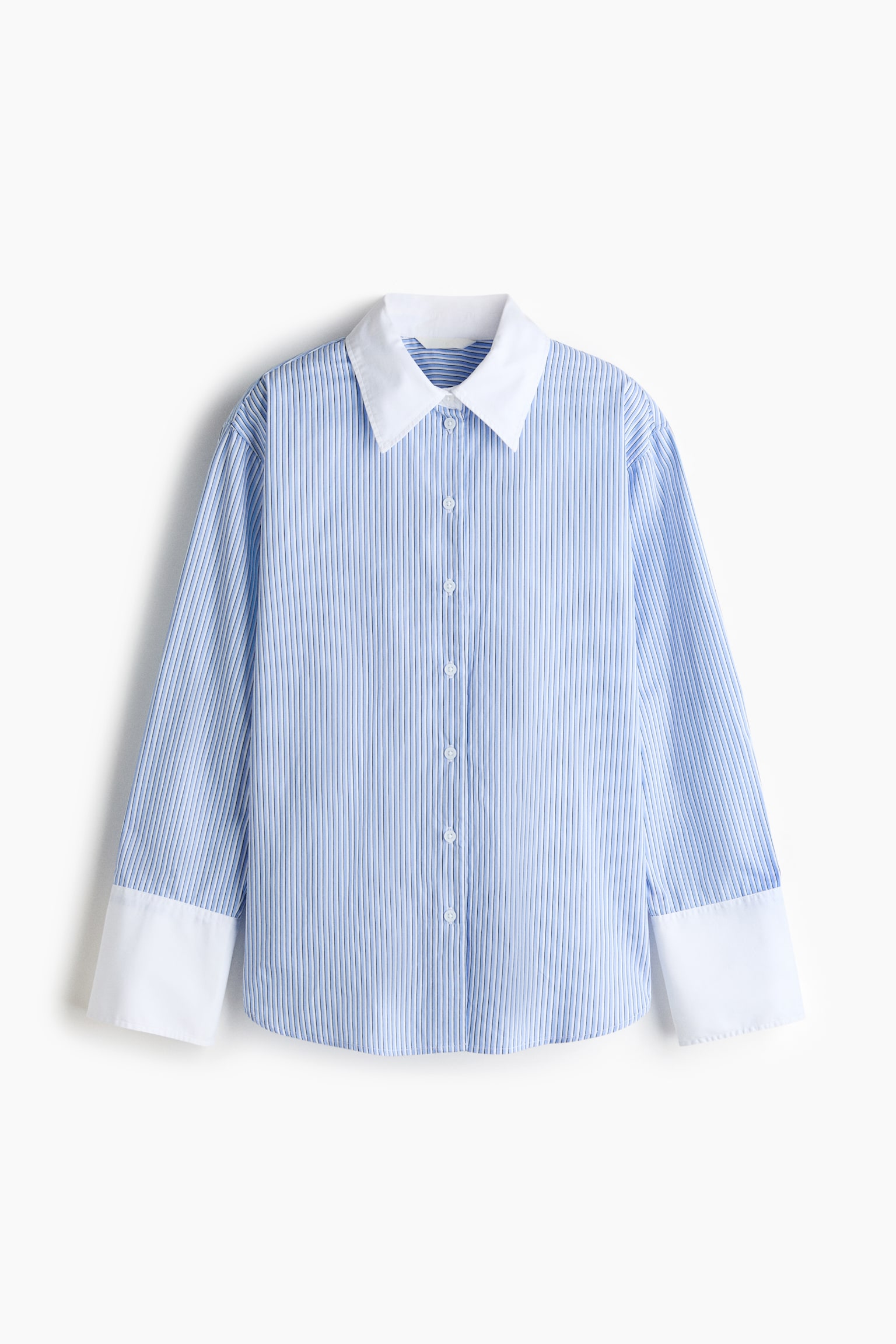 La chemise oversize est présentée de face, arborant un motif à rayures verticales bleues et blanches avec une texture côtelée fine, un col en pointe blanc contrastant, et une patte de boutonnage avant avec des boutons blancs. Elle présente des épaules tombantes, des manches longues avec de larges poignets boutonnés blancs contrastants, et un ourlet droit. - 1