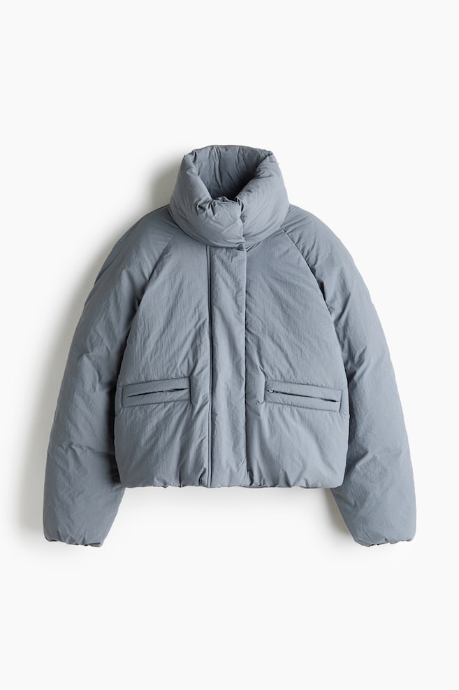 The front-facing cropped puffer jacket is featuring a high, padded stand collar, a hidden front zipper closure, and raglan sleeves. It is displaying a matte slate-grey finish and two horizontal zippered welt pockets on the lower front. - 1
