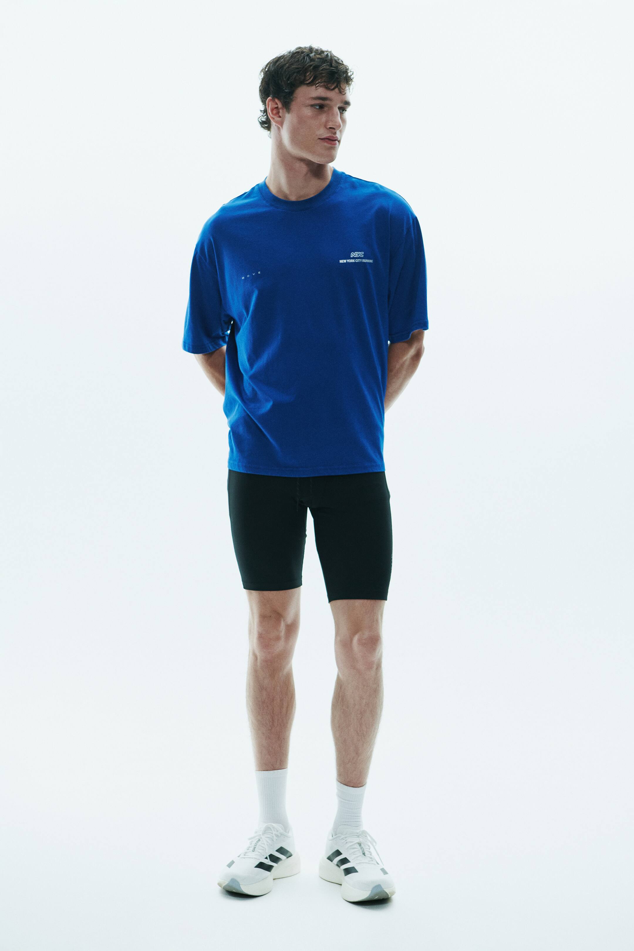 View larger image: A bright blue, short-sleeved crewneck t-shirt featuring small white text on the chest is being worn by the model. The t-shirt is paired with tight-fitting black athletic shorts, white mid-calf socks, and white running sneakers.