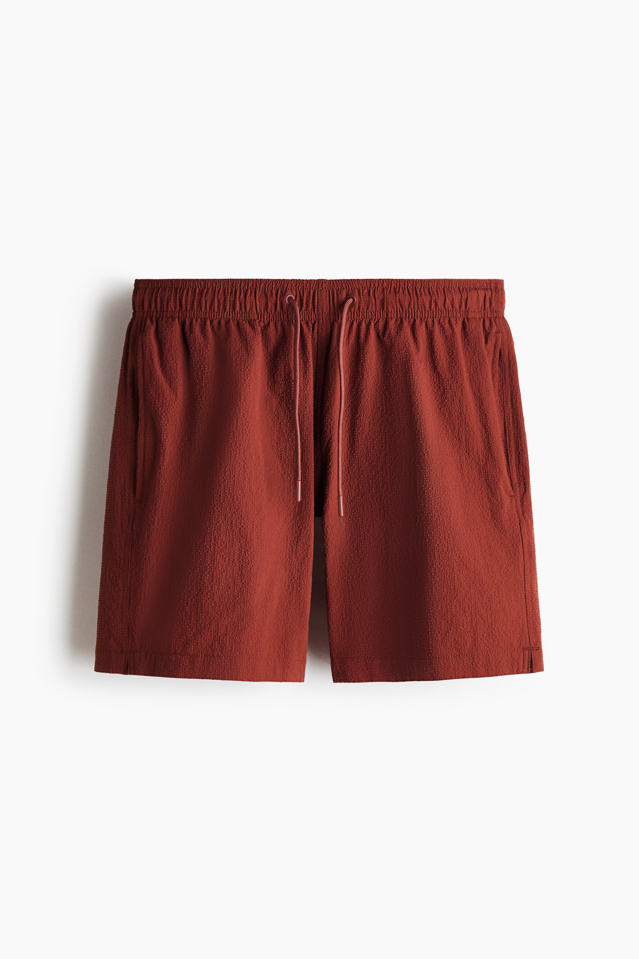 View larger image: The rust-colored shorts feature a textured fabric and an elasticized waistband with a light pink drawstring, presented from the FRONT. They include side pockets and small slits at the hem.
