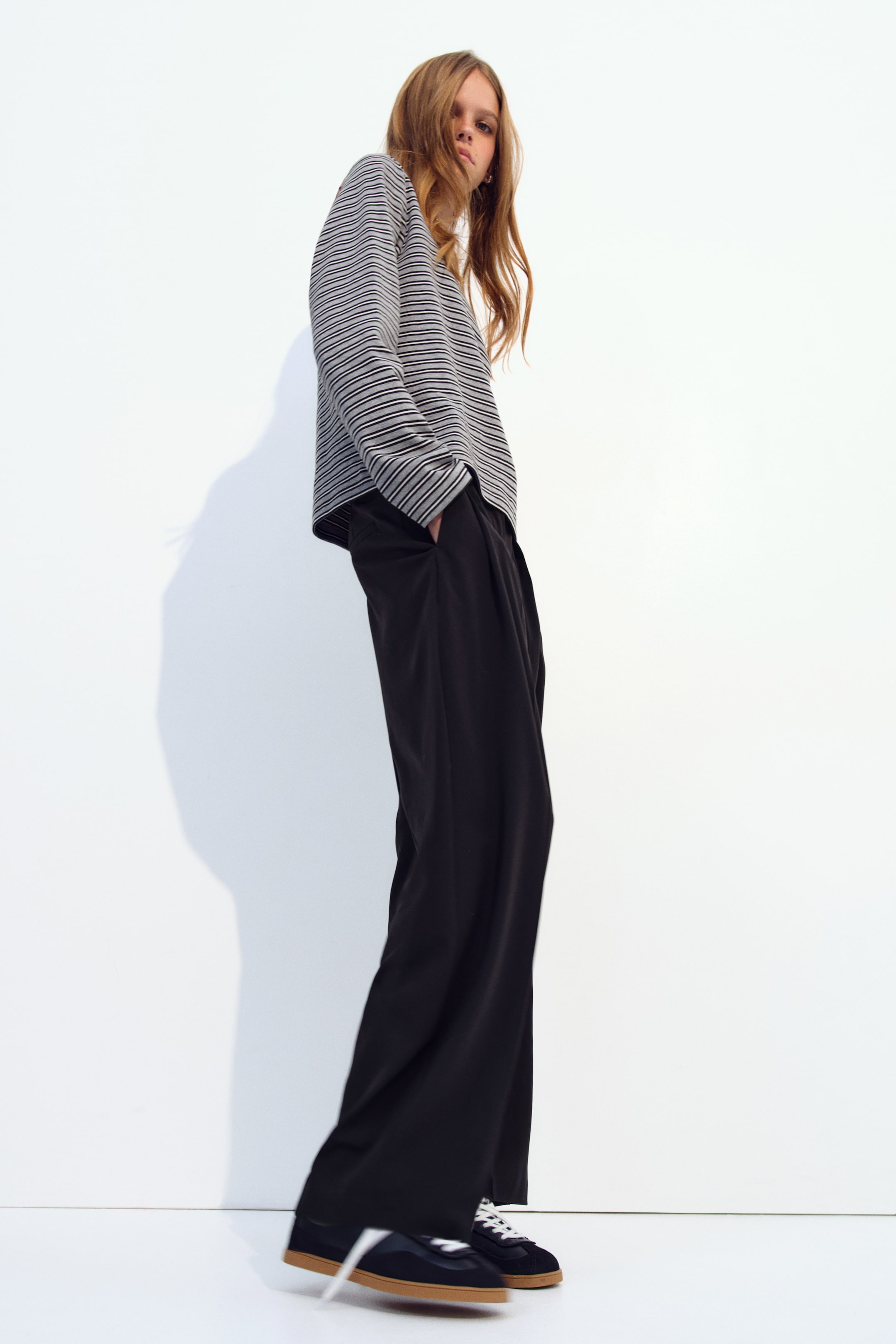 View larger image: Black woven trousers featuring a high waist, front pleats, and a wide barrel-leg cut are worn by the model, who is standing with one hand in a side pocket. The trousers are styled with a striped long-sleeve top and dark trainers.