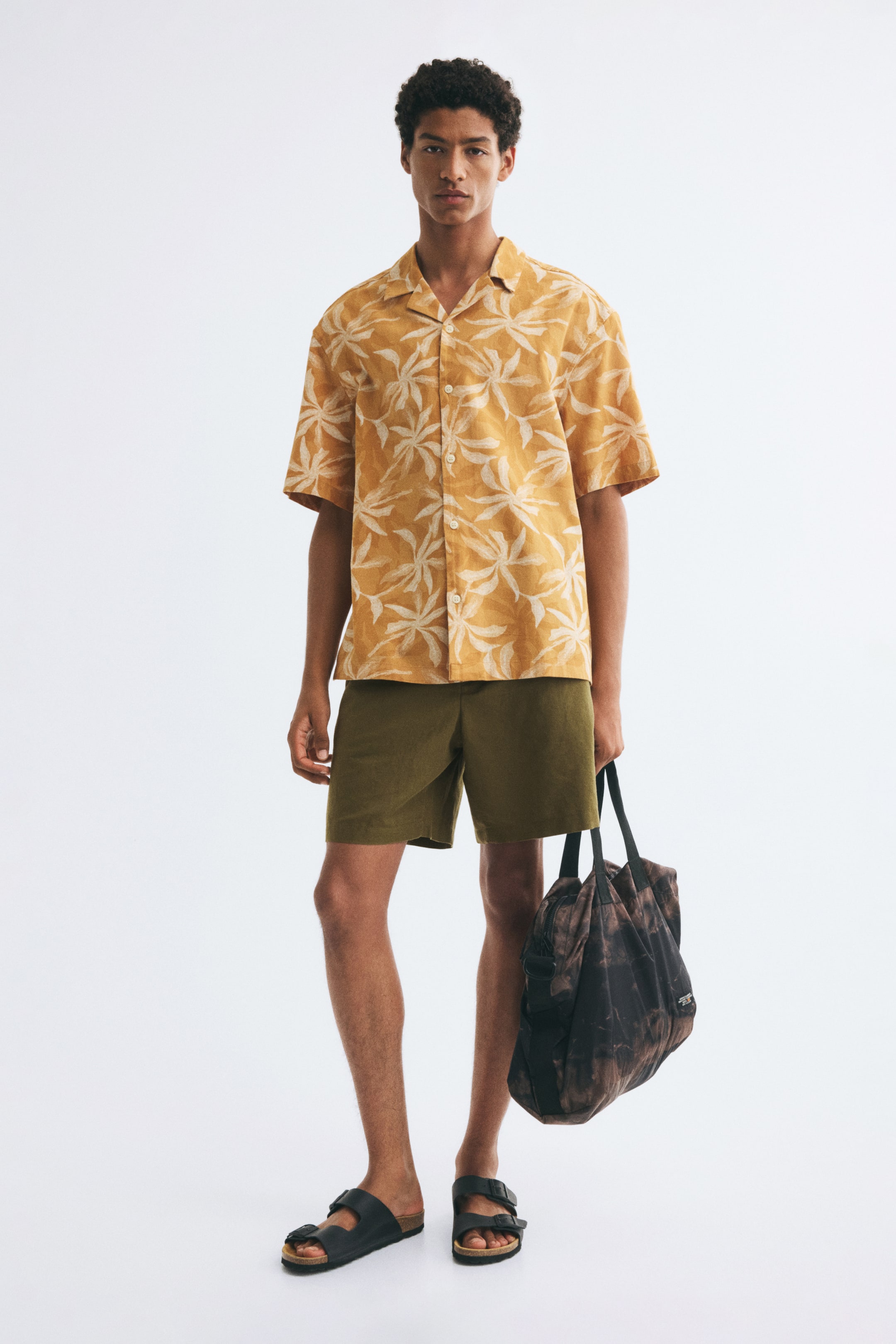 View larger image: The model is wearing olive green shorts made from a textured cotton-linen weave, featuring a regular fit and mid-thigh length. The shorts are styled with a short-sleeved patterned yellow shirt and black slide sandals, while the model holds a dark patterned bag.