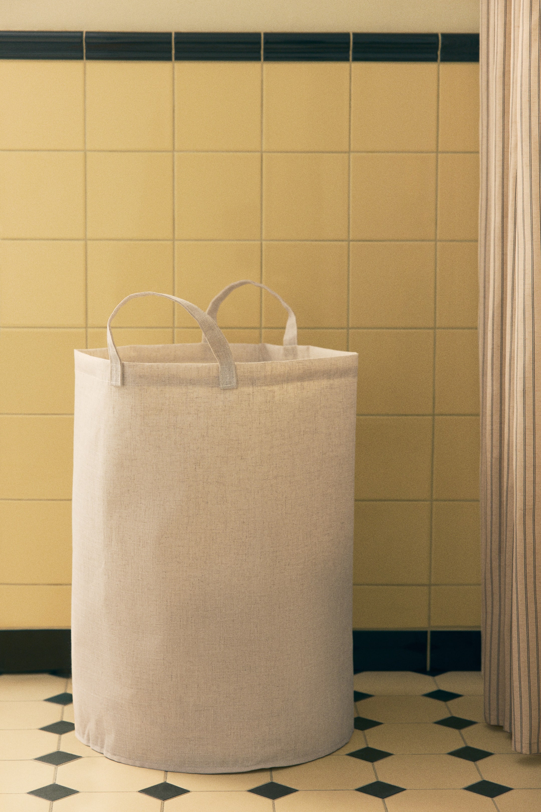 View larger image: A cylindrical laundry bag made from a textured beige linen-blend fabric stands upright, featuring two matching fabric handles at the top rim.