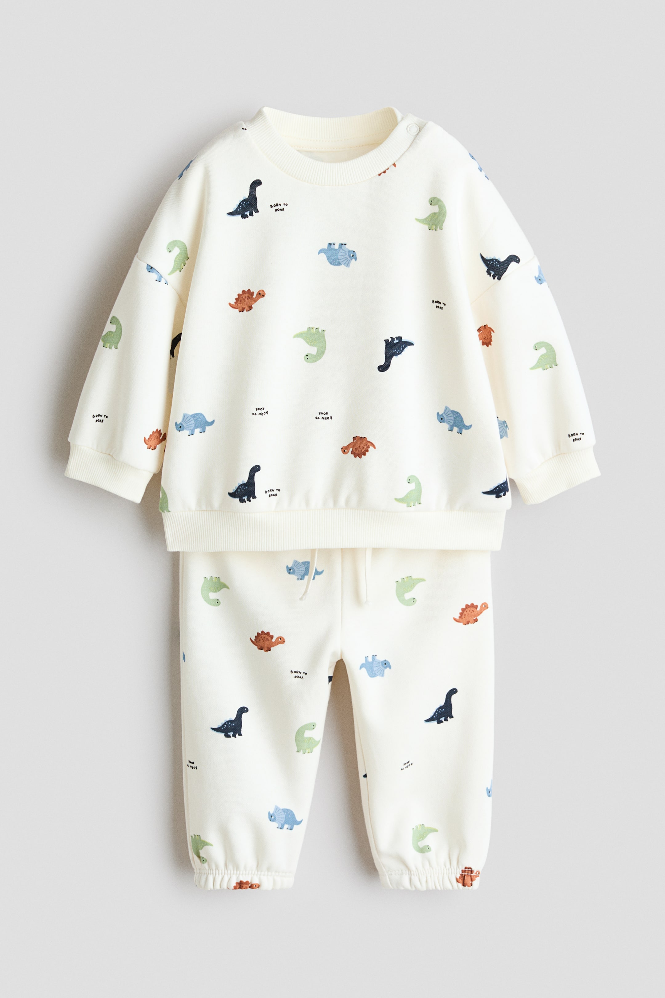 Grotere afbeelding bekijken: Een off-white tweedelig trainingspak voor baby's met een kleurrijke all-over dinosaurusprint. De sweatshirt heeft een ronde hals met schouderknopen, geribde boorden en een geribde zoom, terwijl de bijpassende joggingbroek is voorzien van een elastische tailleband met trekkoord en elastische enkels.