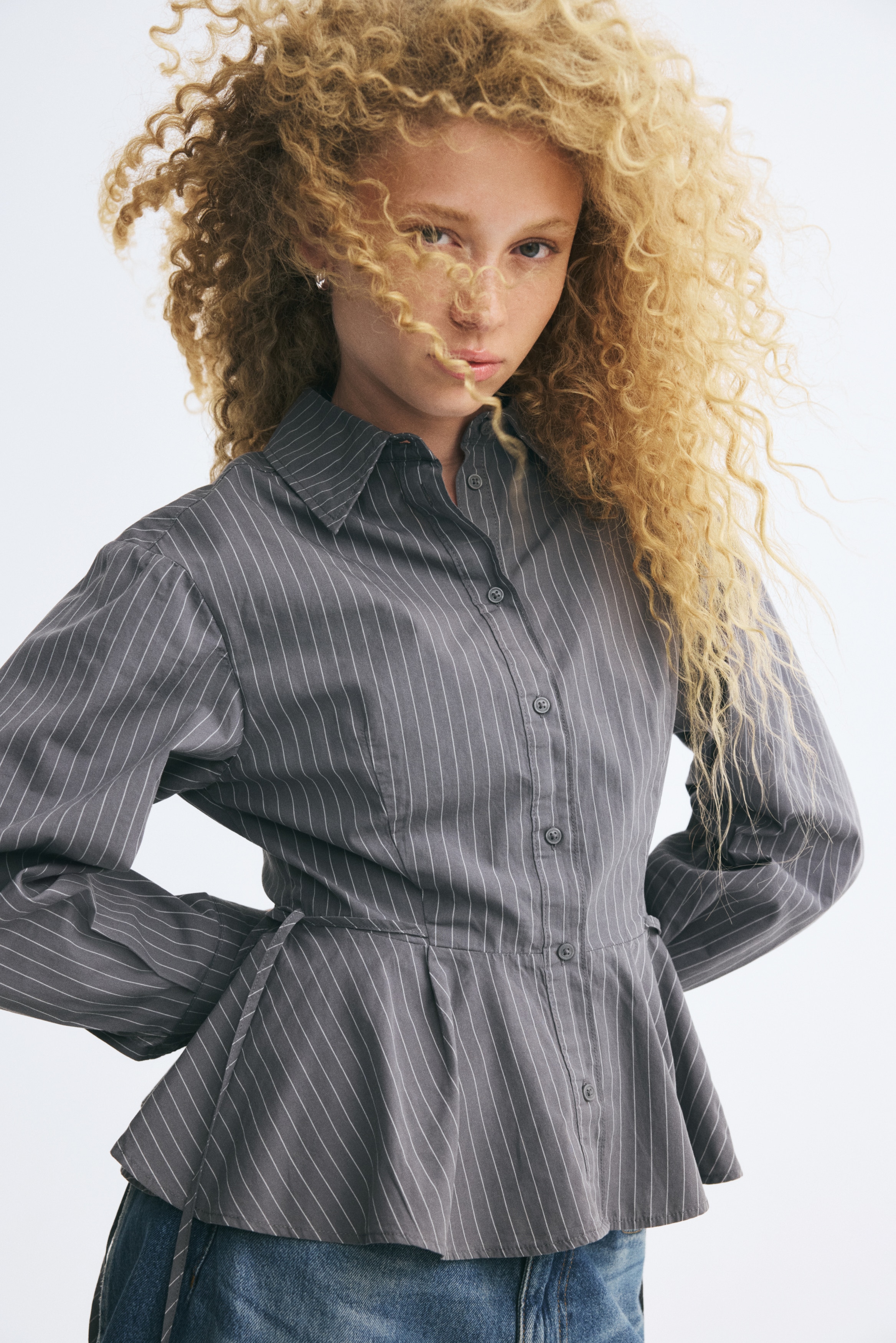 A grey pinstriped shirt is shown facing front, featuring a point collar, a full button-down front, and long sleeves with wide cuffs. The shirt is cinched at the waist with adjustable drawstring side ties that flow into a gathered peplum hem.