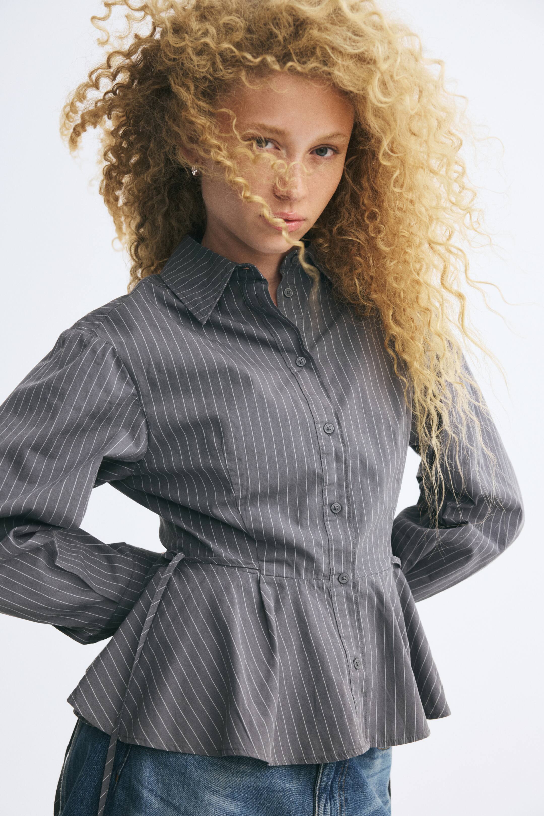 View larger image: The model is wearing a grey, pinstriped button-up shirt with long sleeves, a pointed collar, and a flared peplum hem, styled with blue denim jeans.