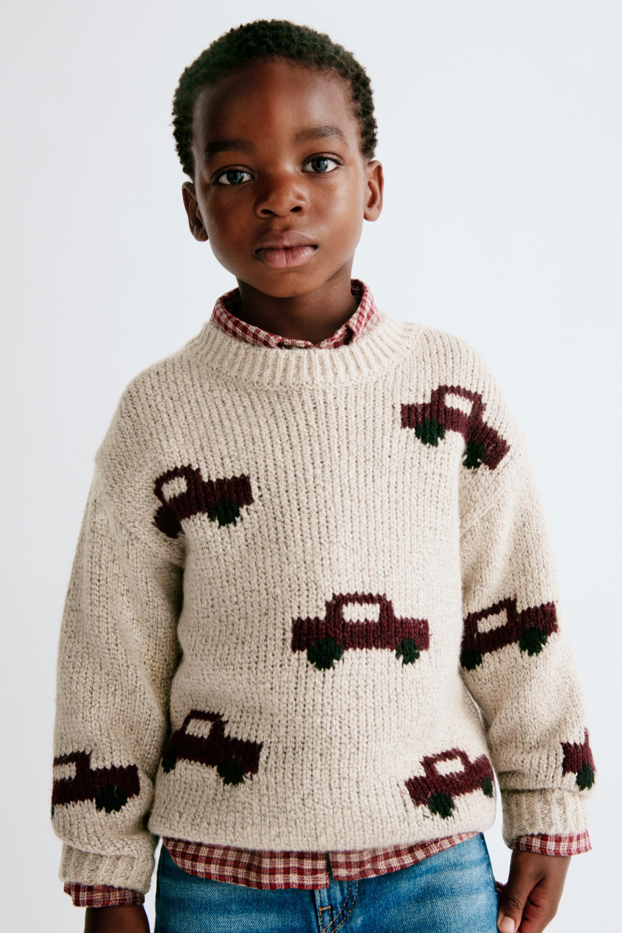 Agrandir l'image: Une vue de face montre un pull en maille jacquard couleur crème, orné d’un motif de voitures bordeaux avec des roues vert foncé. Porté par-dessus une chemise à carreaux rouges et blancs, le pull présente un col rond côtelé, des épaules tombantes ainsi que des finitions côtelées aux poignets et à l’ourlet.