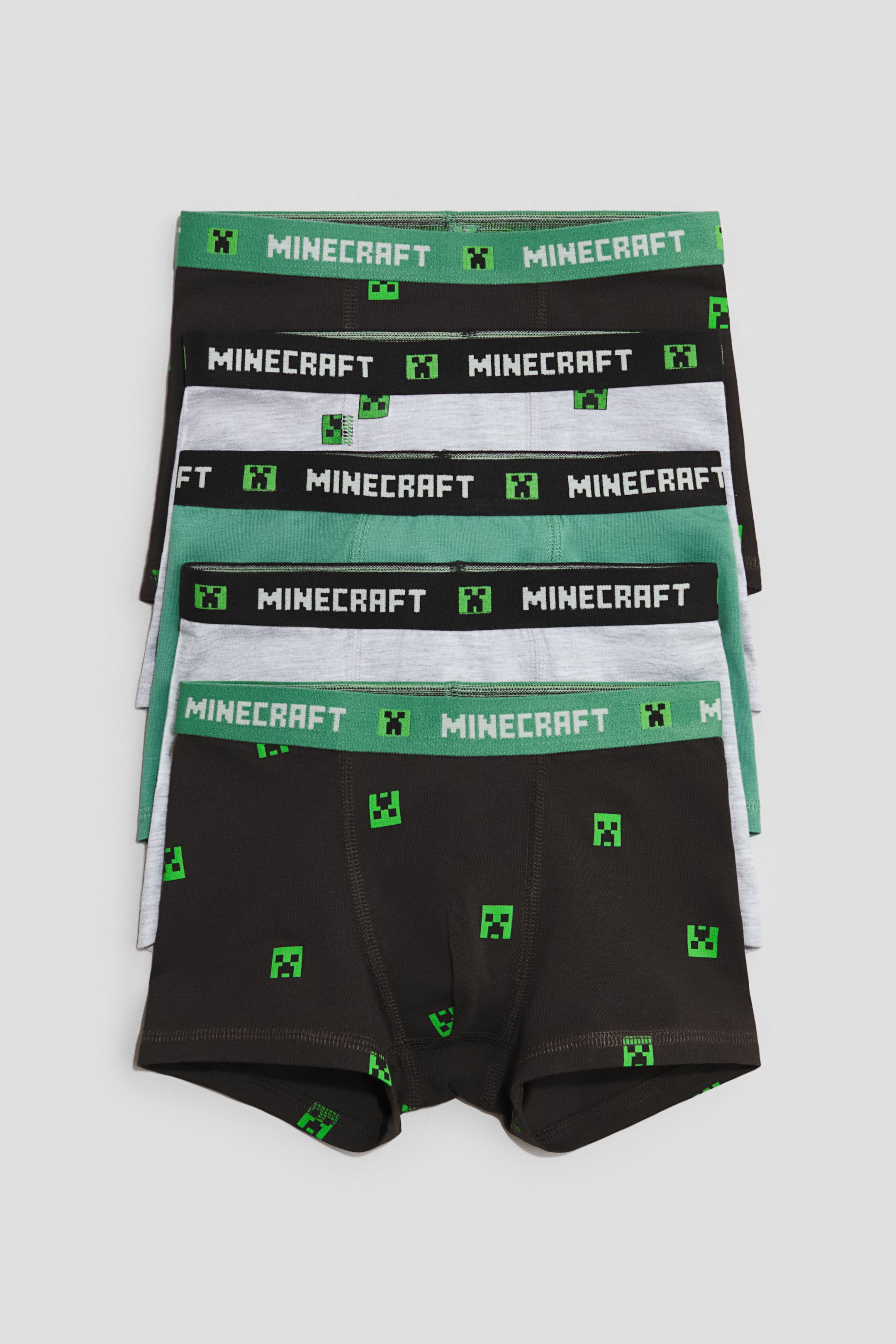 View larger image: A stack of five pairs of cotton jersey trunks is presenting their front views, each featuring short legs, an elasticated waistband, and a lined front. The waistbands are displaying "MINECRAFT" text and pixelated green creeper faces, while the trunks are alternating in color between black with green creeper patterns, solid green, and grey with green creeper patterns.