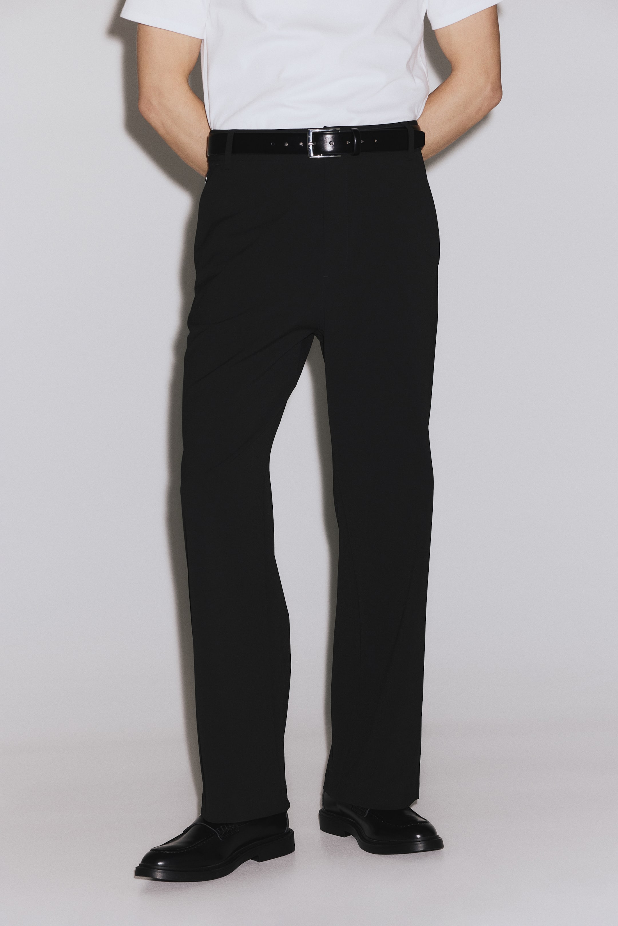 Ampliar la imagen: Los pantalones negros presentan un corte recto a lo largo de la pierna, mostrando una tela lisa sin arrugas. Un cinturón negro sujeta la cintura, y el dobladillo del pantalón descansa sobre zapatos negros.
