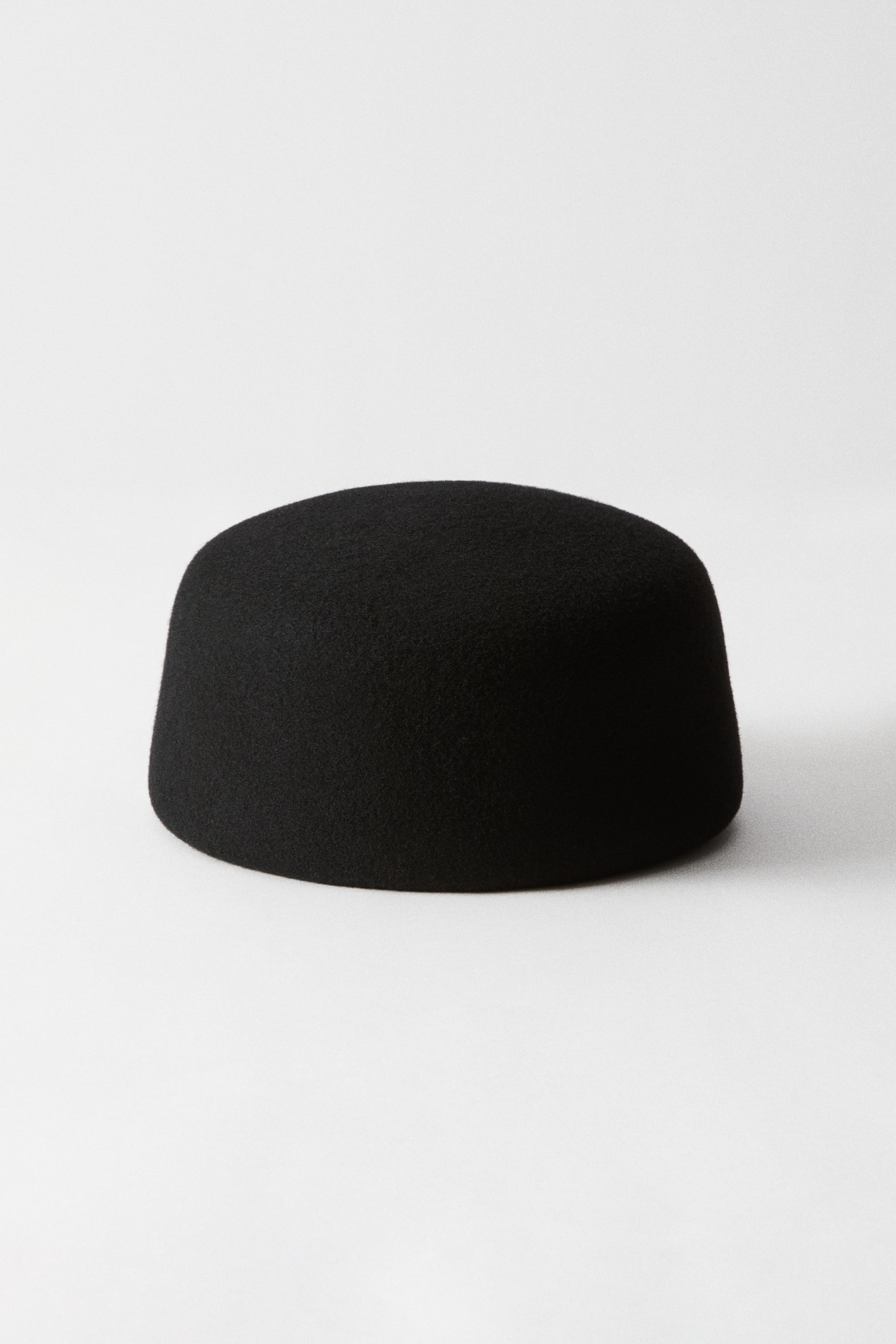 Ver imagen más grande: Un sombrero pillbox negro, confeccionado en lana afieltrada, muestra su diseño cilíndrico estructurado, su copa plana y su acabado mate sin costuras desde un ángulo frontal.