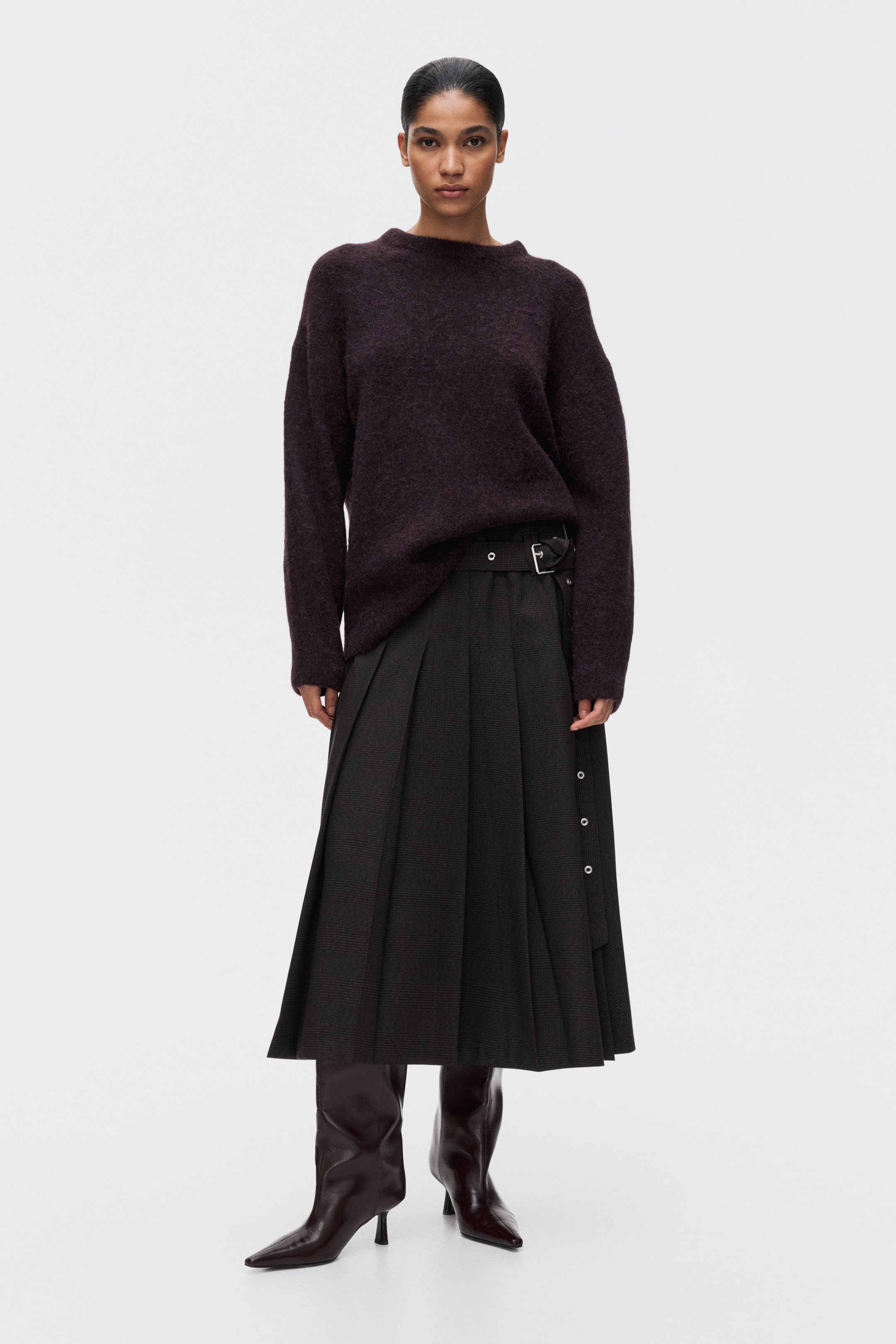 Ampliar la imagen: Un suéter de cuello redondo púrpura oscuro con una textura peluda se está usando, combinado con una falda midi plisada de color gris oscuro. La falda presenta un cinturón negro con una hebilla plateada y una línea de botones a presión plateados en su lado derecho, que se extiende sobre unas botas puntiagudas de color marrón oscuro.
