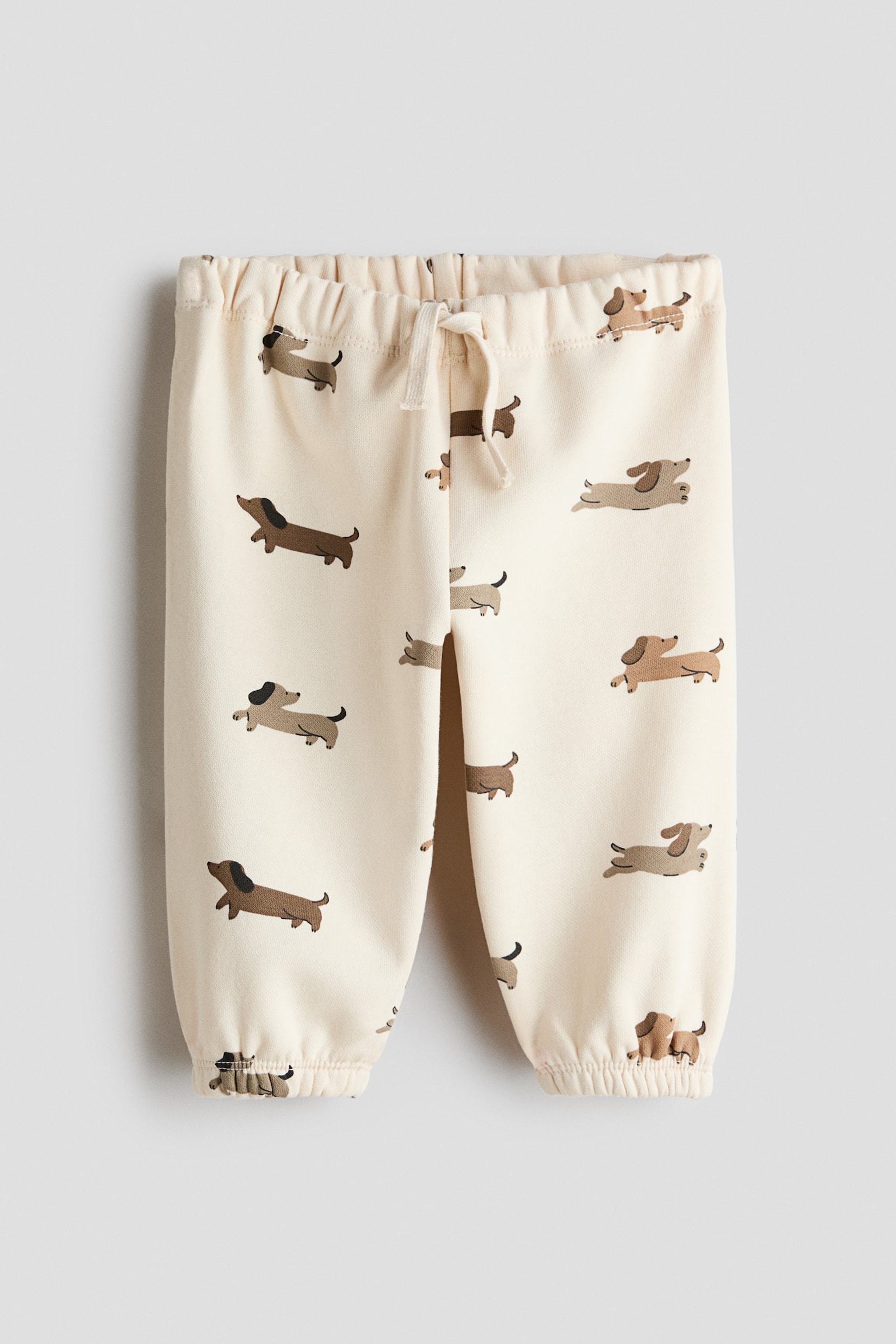 A pair of cream-colored track pants is shown from the front, featuring an all-over print of dachshund dogs in shades of brown. The design includes a gathered elastic waistband with a decorative cream drawstring tied at the center and gathered elastic cuffs at the hems.