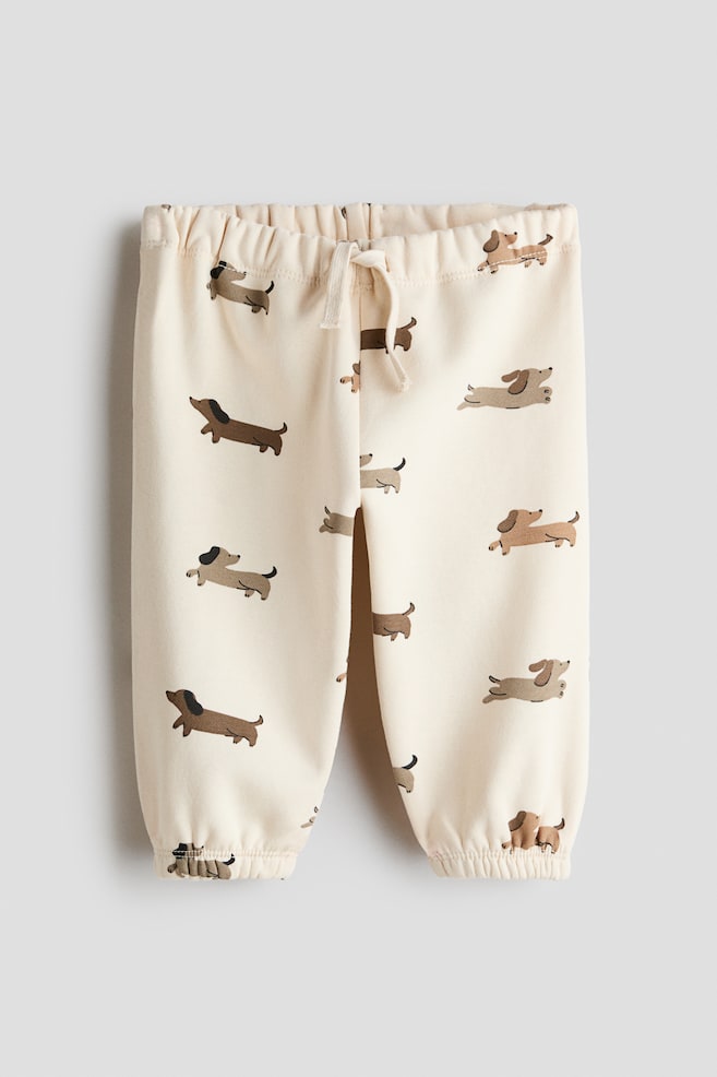 A pair of cream-coloured track pants is shown from the front, featuring an all-over print of dachshund dogs in shades of brown. The design includes a gathered elasticated waistband with a decorative cream drawstring tied at the centre and gathered elasticated cuffs at the hems. - 1