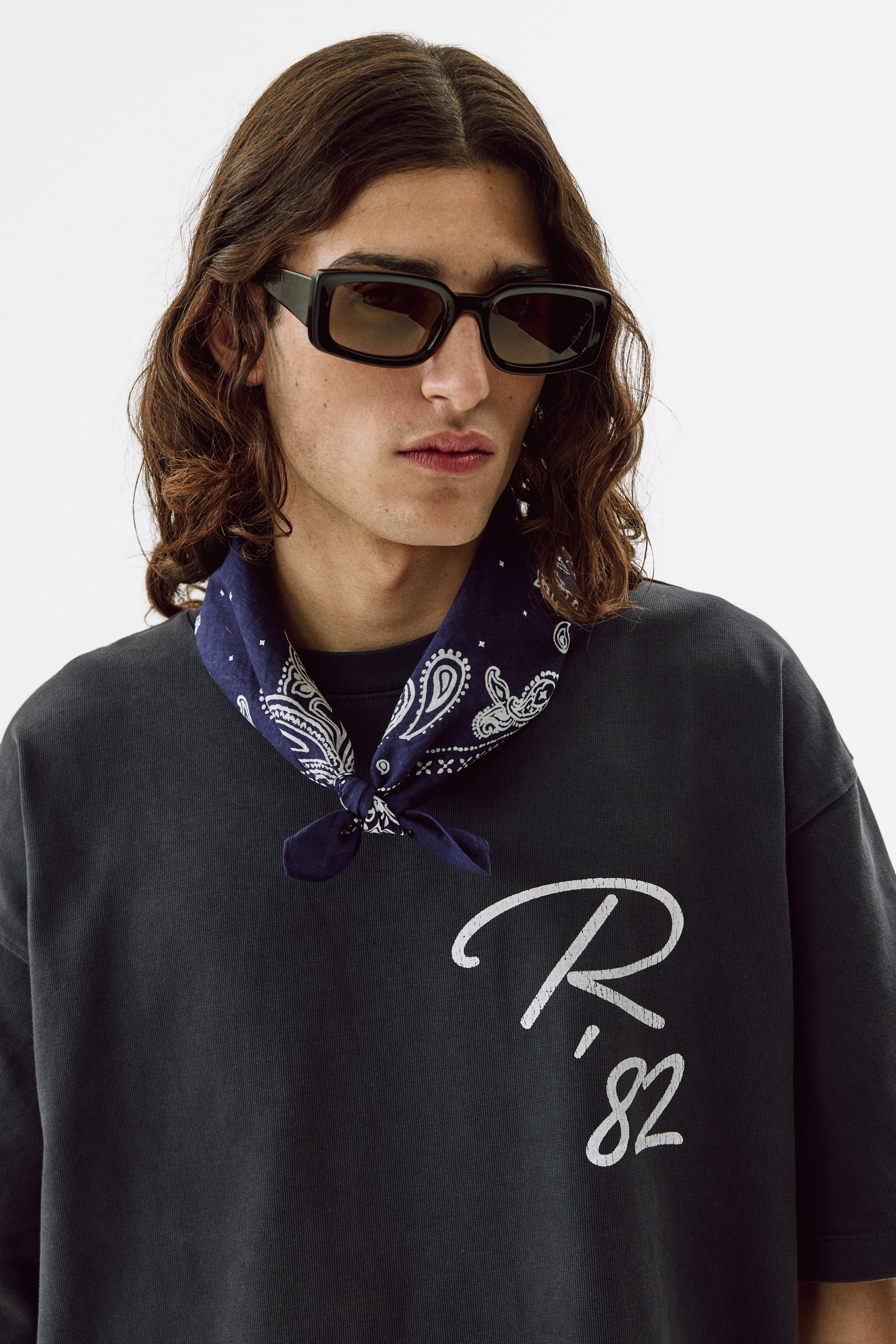 View larger image: The model is wearing a dark gray crew neck t-shirt, showcasing a white distressed graphic of a script 'R' and ',82' on the front. A blue paisley bandana is tied around the neck, resting on the neckline.