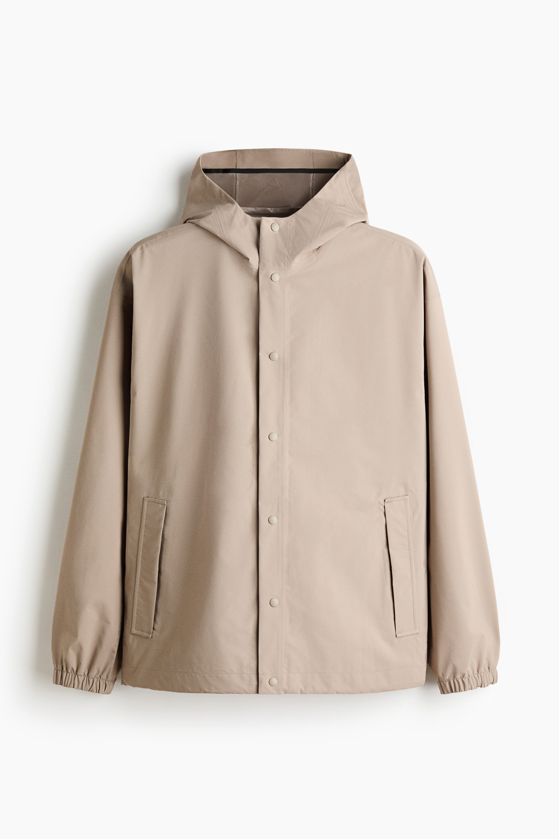 Ver imagen más grande: Una chaqueta beige se presenta desde el frente, con una capucha incorporada que muestra un forro de malla oscura visible dentro del cuello, un cierre frontal completo con botones de presión, dos bolsillos rectangulares con solapa en la parte inferior delantera, y puños y dobladillo elásticos.