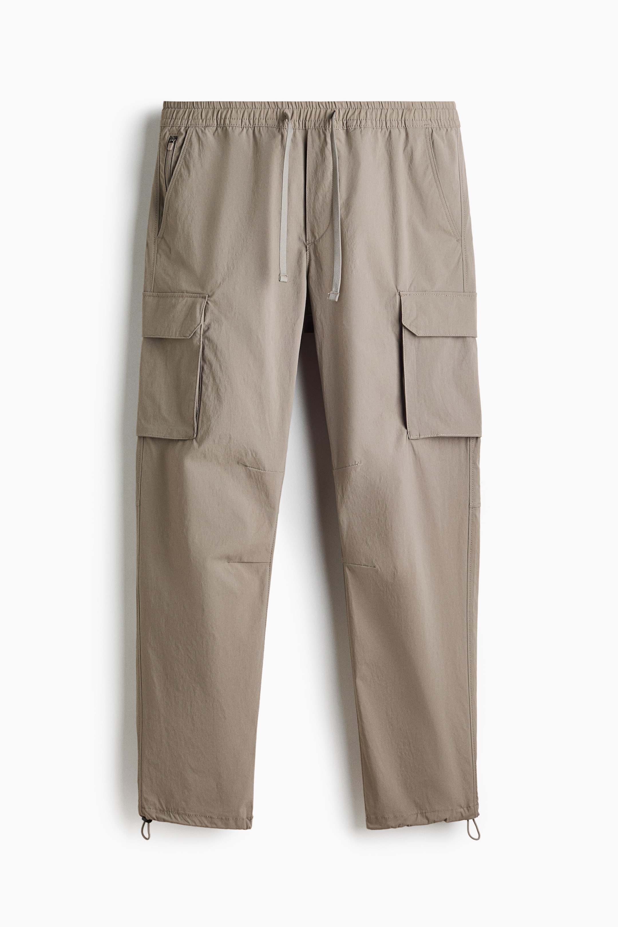 View larger image: Taupe cargo pants are showcased in a front view, highlighting an elasticated drawstring waistband and two front slash pockets, one with a zipper closure. Two large flap cargo pockets are positioned on the outer thighs, and adjustable drawstrings are evident at the ankle cuffs.