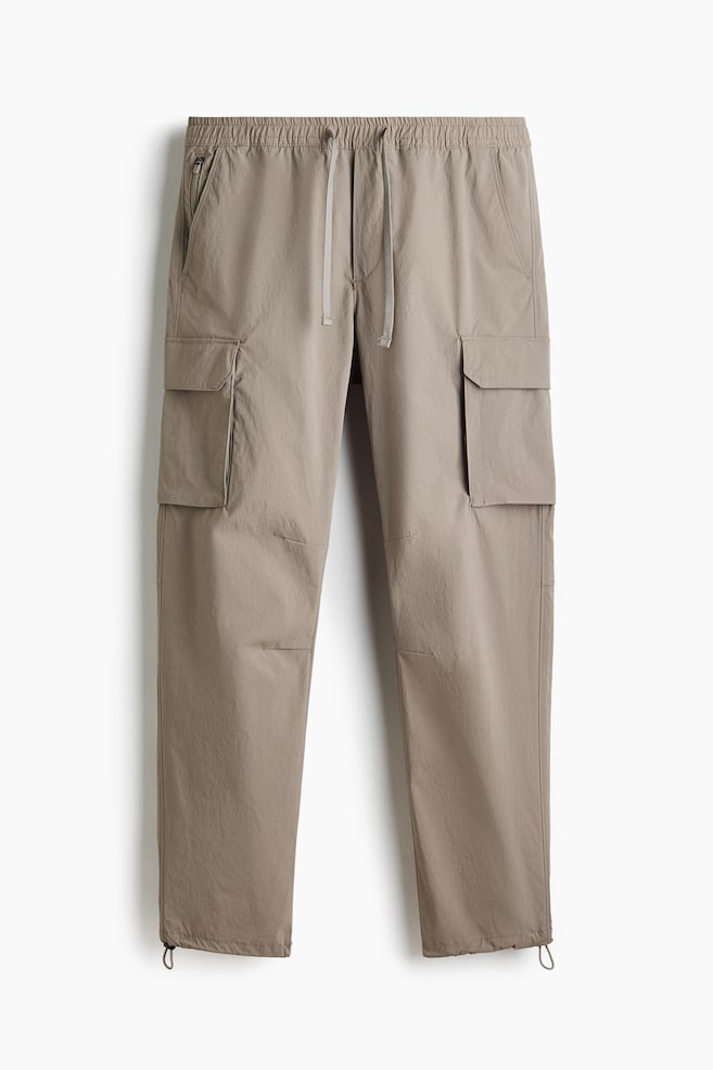 Taupe cargo pants are presenting a front view, featuring an elasticated drawstring waistband and two front slash pockets, one displaying a zipper closure. Two large flap cargo pockets are appearing on the outer thighs, and adjustable drawstrings are visible at the ankle cuffs. - 1