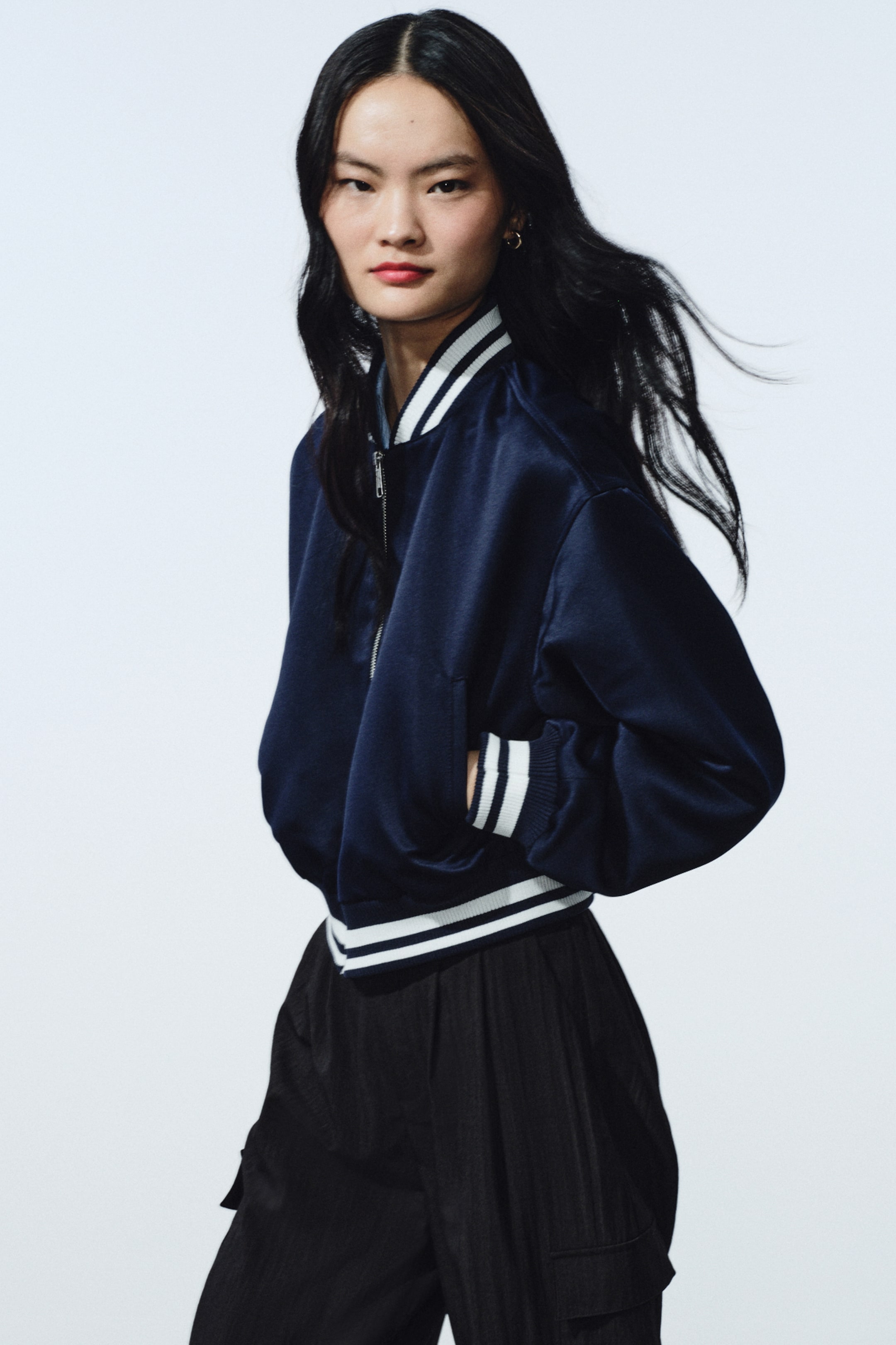 拡大画像を表示: サテンのような光沢を持つネイビーブルーのボンバージャケットを、やや左向きに配置して撮影しています。フロントはジッパー開閉で、白とネイビーのストライプ入りリブの襟・袖口・裾を備え、サイドポケットに手を入れた状態です。