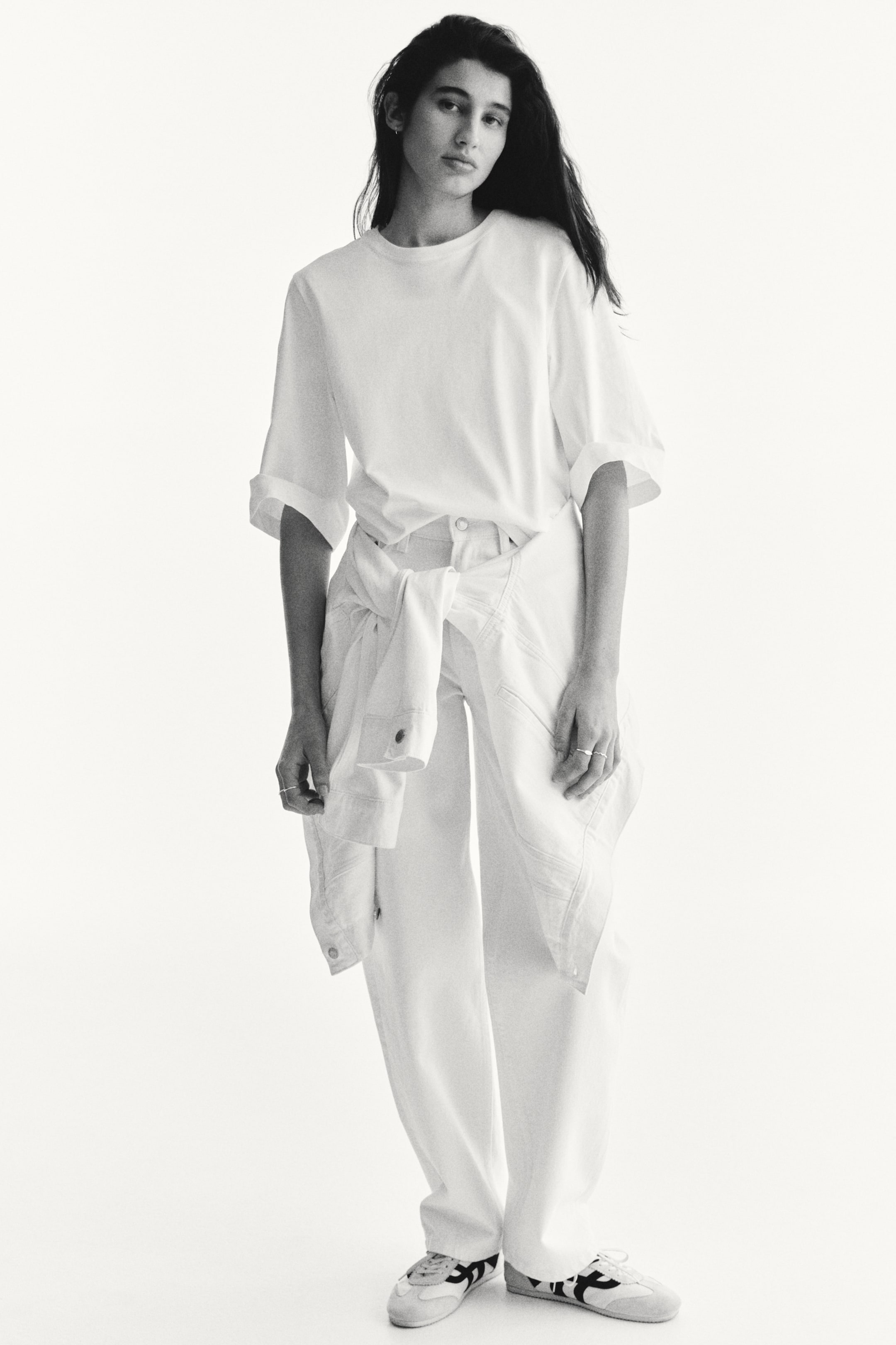 查看大圖: 白色寬鬆版型T恤,特色是捲起的短袖和圓領,搭配寬鬆的白色長褲,顯露出明顯的縫線細節。腰間系著一件白色長袖襯衫,完成了這套單色造型,搭配淺色運動鞋,運動鞋上有深色對比條紋。