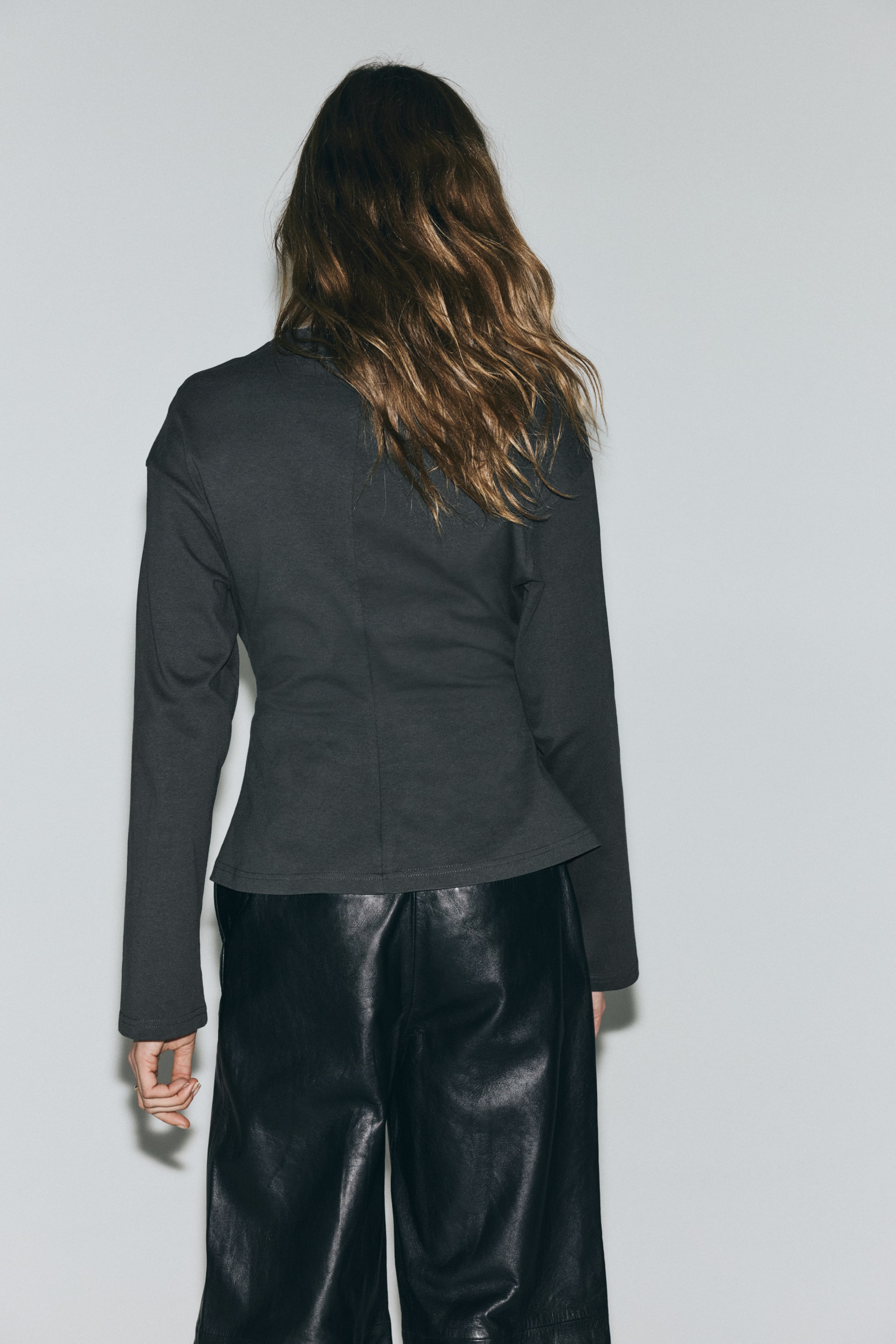 Преглед на по-голямо изображение: ГЪРБЪТ на тъмно сиво горнище е на показ, с паднали рамене и дълги ръкави. То демонстрира прилепнала талия, подчертана от вертикални шевове, които след това се разширяват в широка подгъвка.