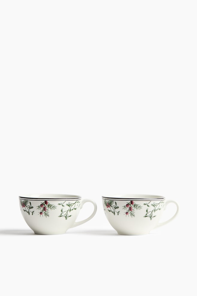 Due tazze in ceramica color bianco sporco sono affiancate, rivolte verso il fronte, e mostrano un motivo botanico con foglie verdi e bacche rosse sotto un sottile bordo scuro vicino al bordo superiore. Manici curvi si estendono dal lato destro di ciascuna tazza. - 1