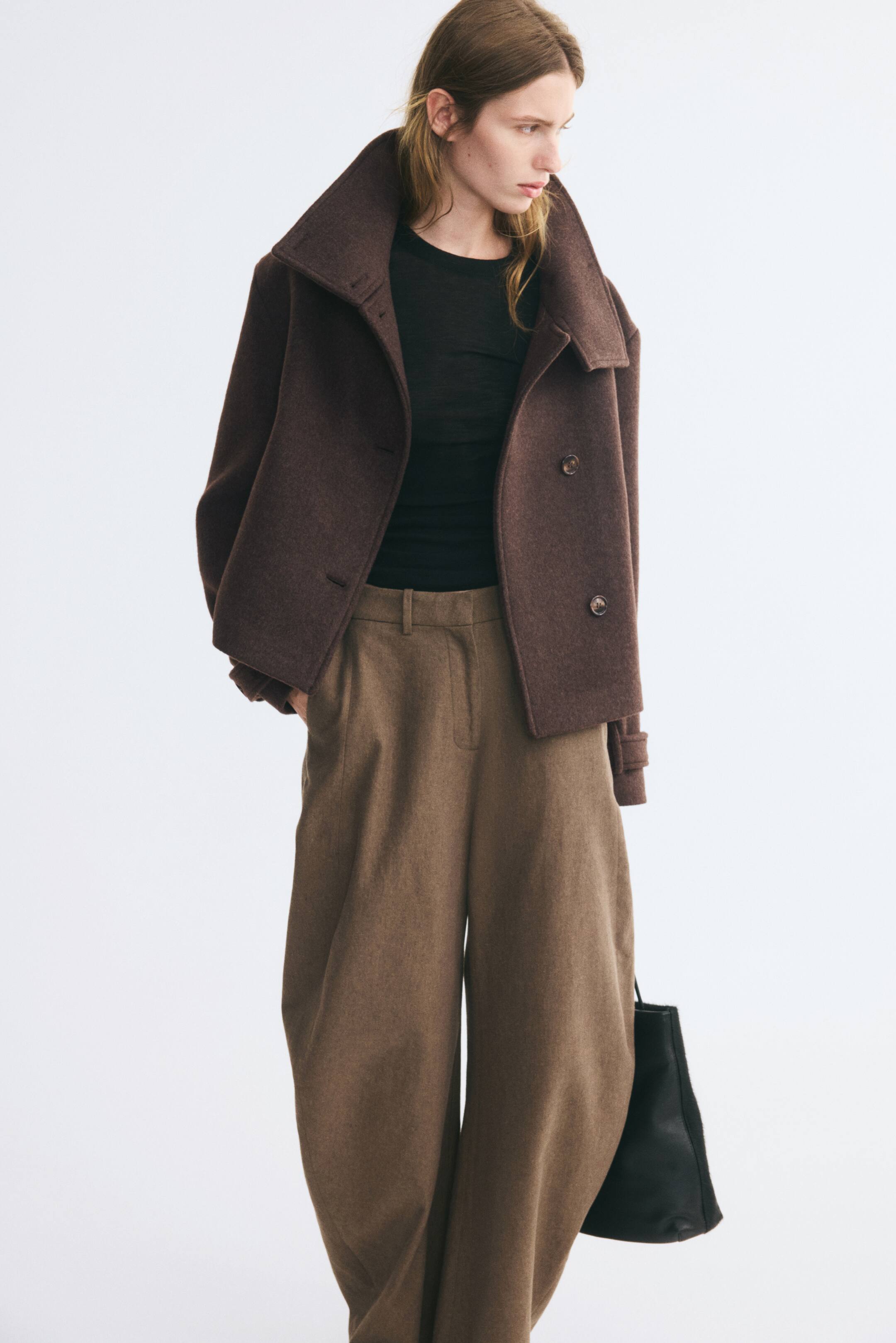 Grotere afbeelding bekijken: Een donkerbruine, oversized jas van een wolmix met een brede, open kraag en knoopdetails aan de voorkant wordt open gedragen door het model. De jas is gelaagd over een aansluitende zwarte top en gestyled met een wijde bruine pantalon.