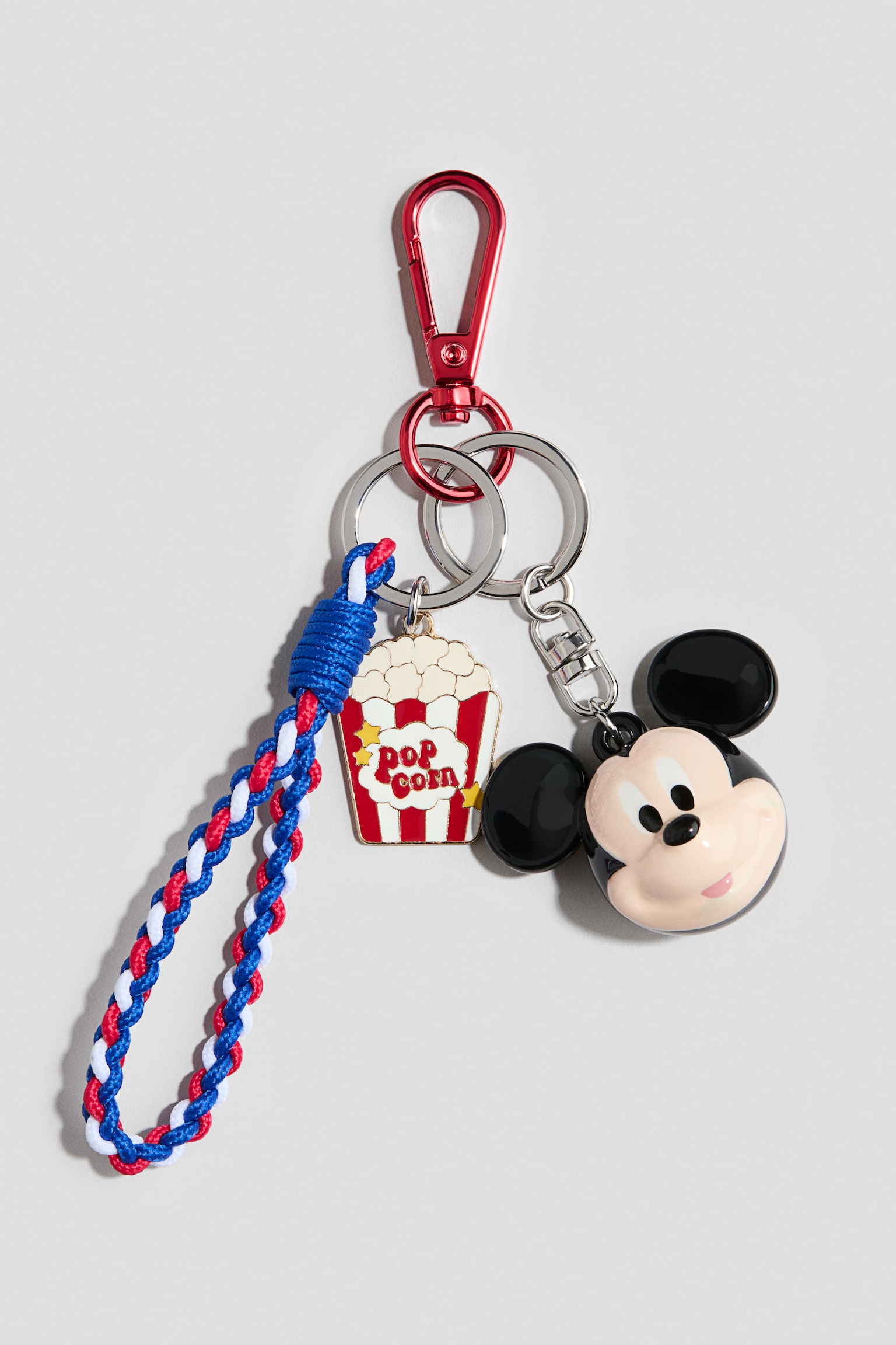 A bag charm is hanging from a red metal carabiner hook connected to silver split rings, showcasing three distinct decorations. A braided red, white, and blue cord loop, a flat metal popcorn bucket charm, and a three-dimensional Mickey Mouse head charm facing front are dangling from the rings.