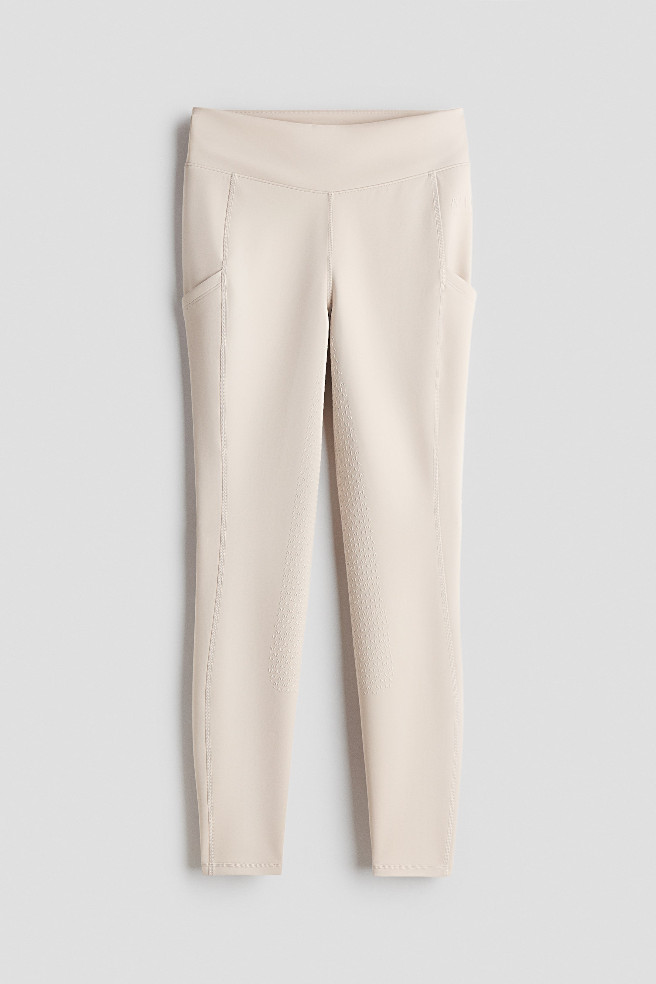View larger image: A pair of light beige riding leggings is shown from the front, featuring a high waist with a wide waistband and side pockets on both thighs. A silicone grip pattern covers the inner leg, and elasticated binding finishes the hems.