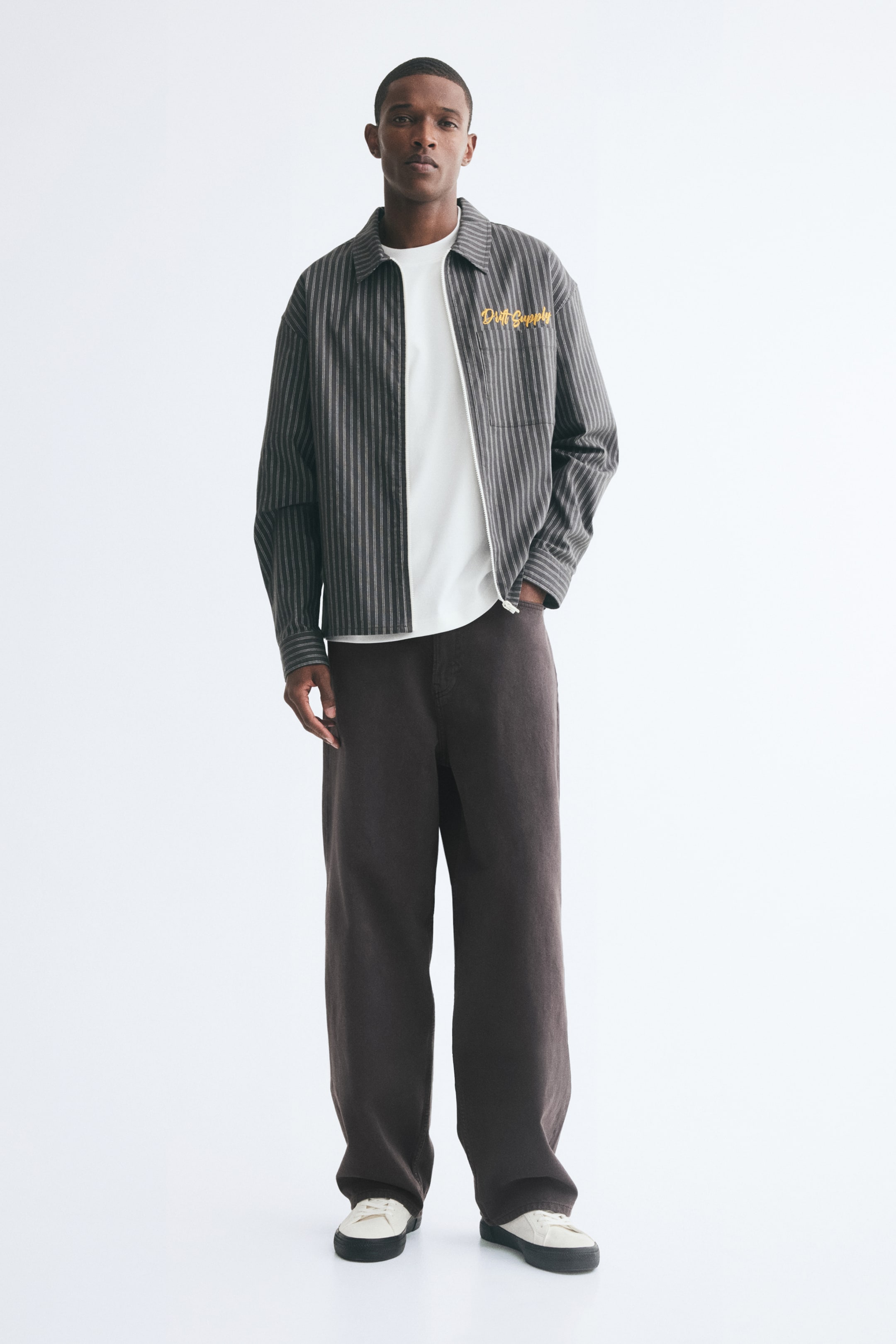 View larger image: Dark brown baggy jeans in sturdy denim are worn by the model, featuring a loose, rounded leg shape that pools slightly at the ankles. The jeans are styled with a white T-shirt, a striped zip-up jacket, and white sneakers, with the model resting one hand in the front pocket.