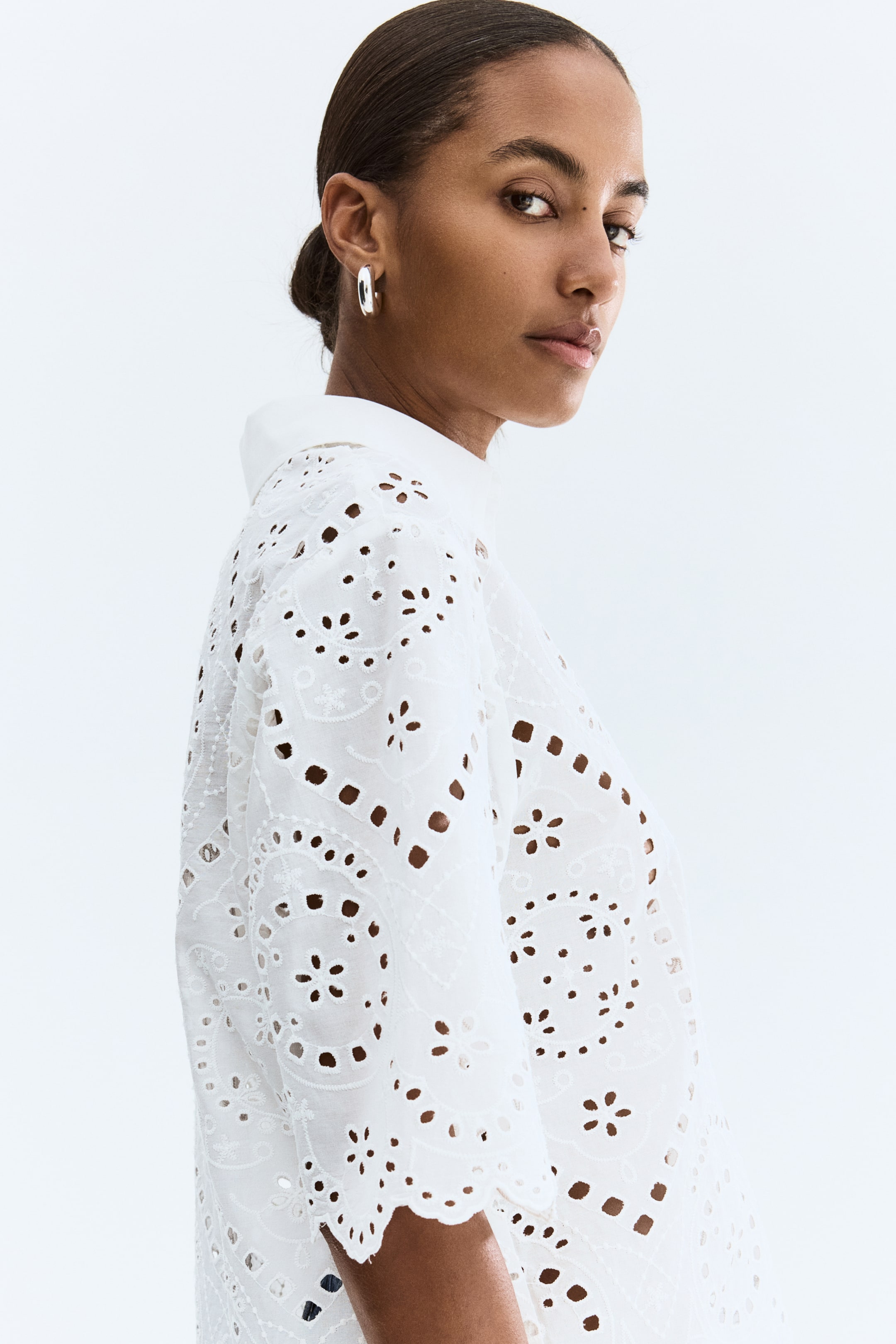 Ampliar la imagen: Una camisa blanca de broderie anglaise muestra patrones intrincados de flores y figuras geométricas recortadas. Presenta un cuello en punta y mangas anchas con bordes festoneados, con la piel visible a través de los ojales, orientada hacia la derecha.
