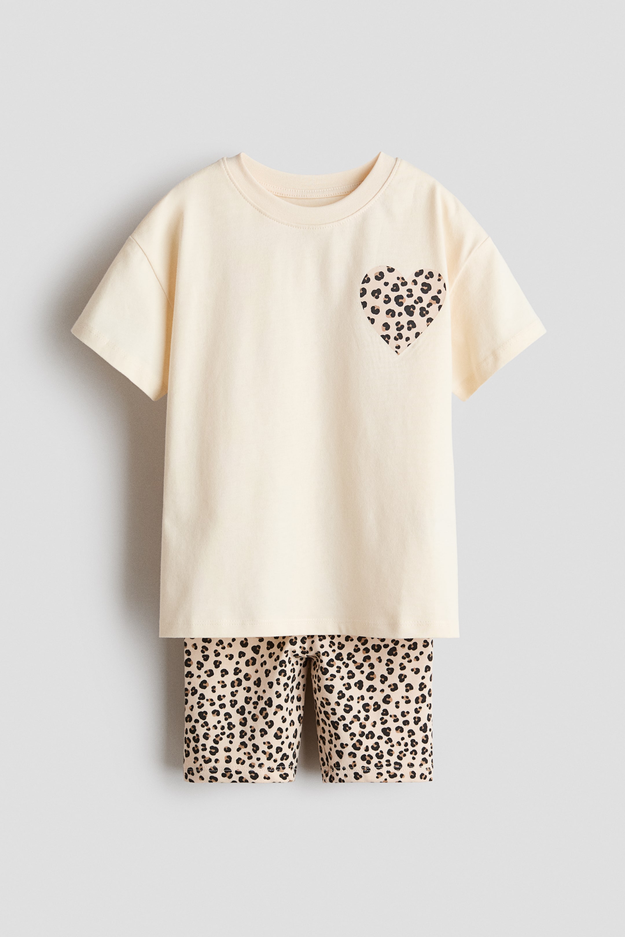 Visa större bild: Ett krämfärgat kortärmat t-shirt och shortsset med leopardmönster visas framifrån. T-shirten har en rund hals och ett hjärtformat leopardmönstrat märke på vänster bröst, medan shortsen visar ett matchande leopardmönster.