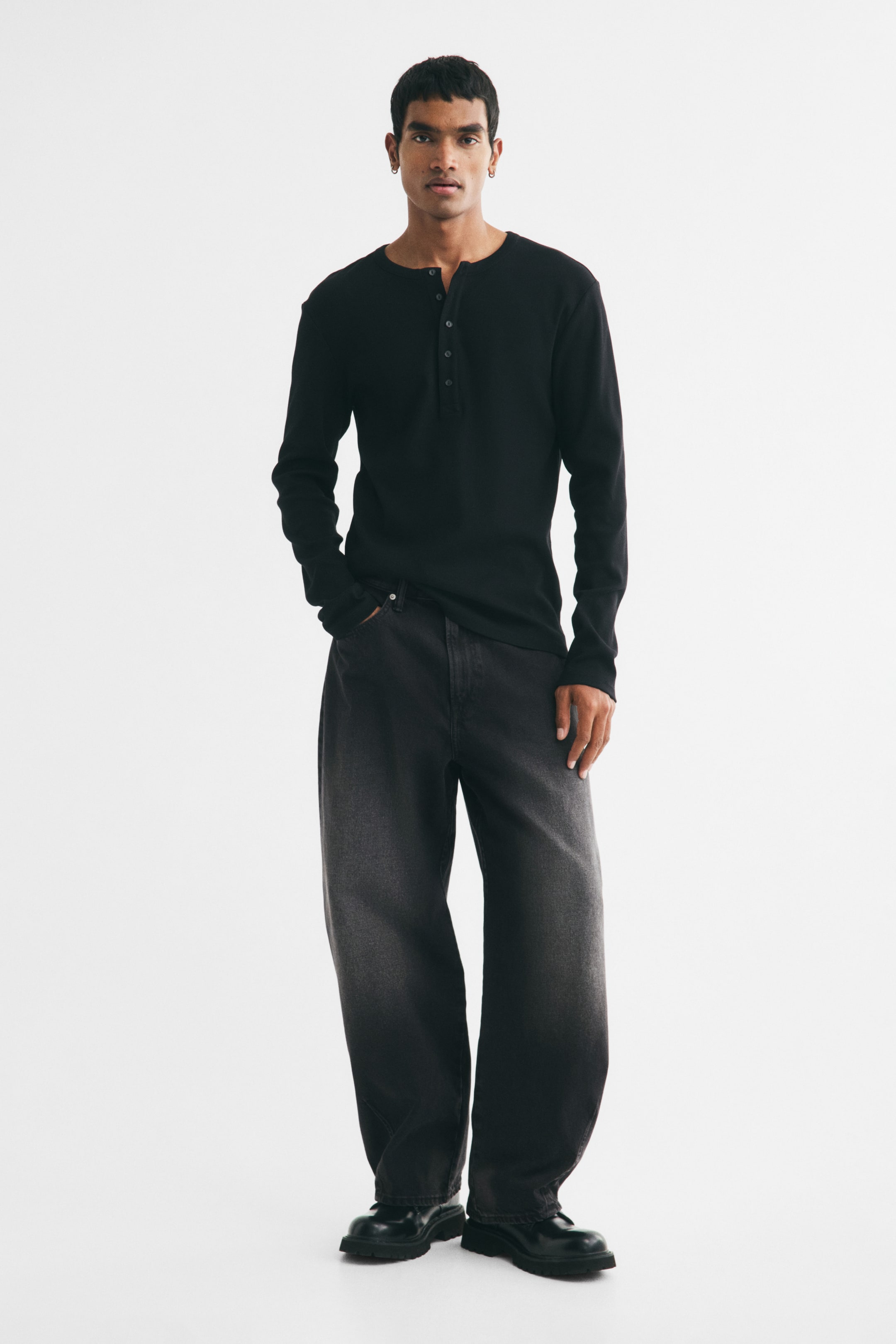 Vis større bilde: En svart henley-skjorte med lange ermer, med stolpe med tre knapper, bæres av modellen og er stylet med mørkvaskede, løstsittende jeans.