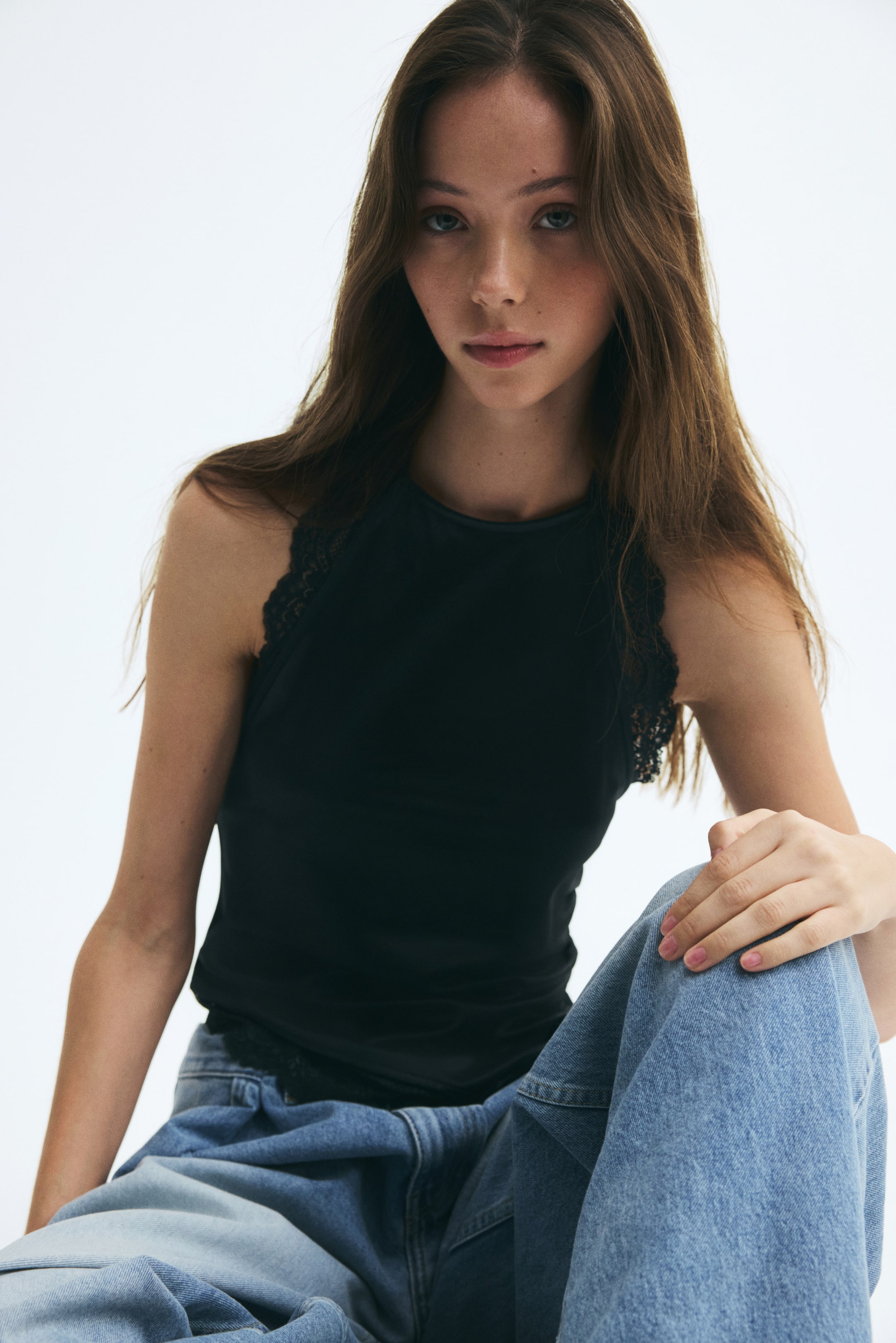 View larger image: A black satin halterneck top with a high round neckline and scalloped lace trim along the armholes is being worn by the model. The top is paired with light-wash blue jeans.