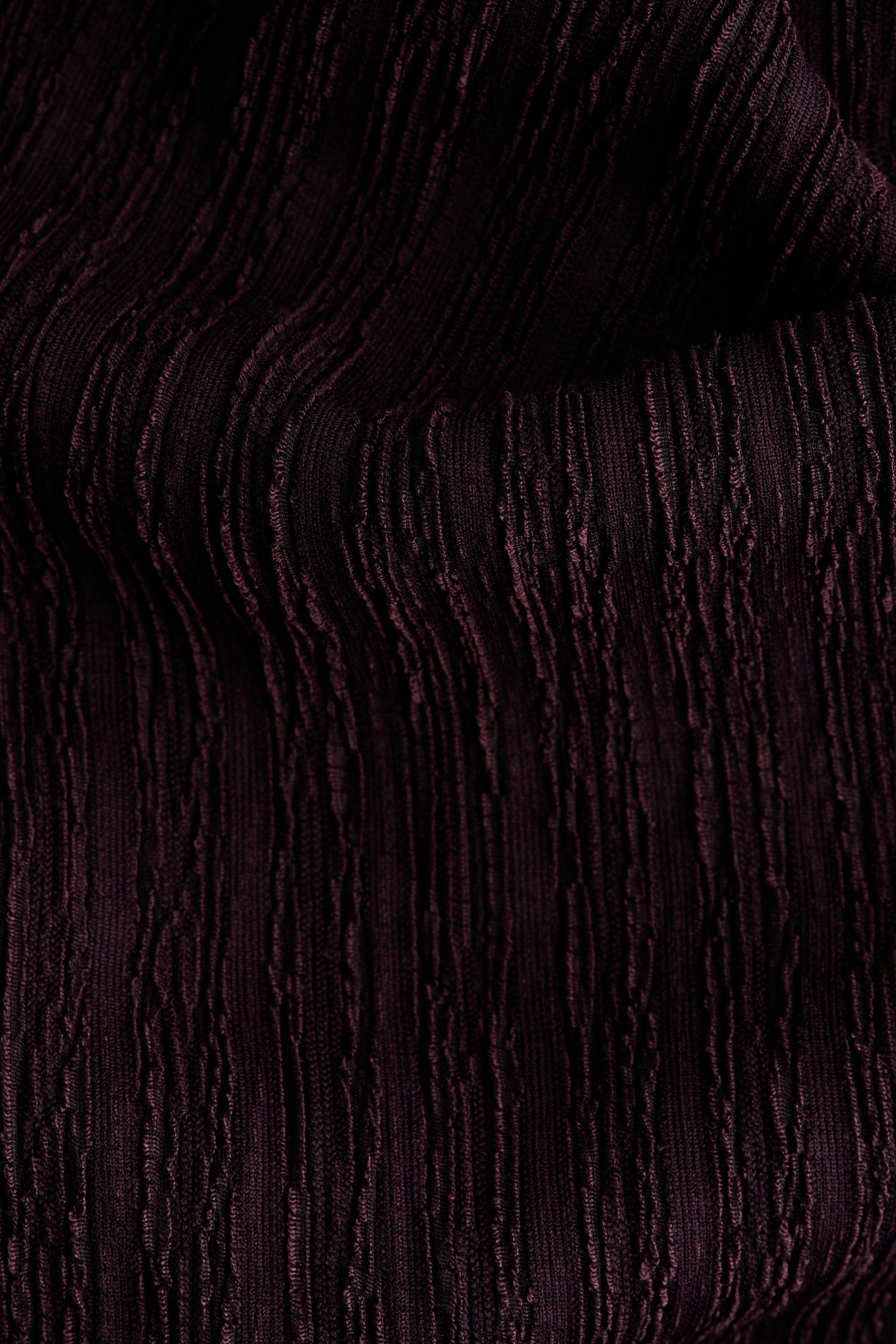 Größeres Bild ansehen: Ein dunkelburgunderfarbener Stoff zeigt eine fein geknitterte Textur mit unregelmäßigen vertikalen Linien und Falten. Er fällt weich und zeigt Tiefe und Schatten.